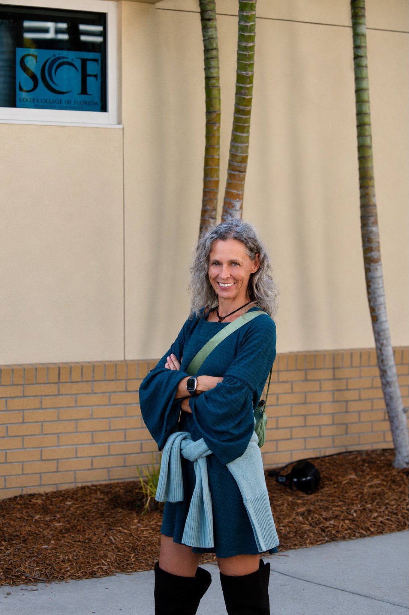 A woman with curly gray hair smiling with arms crossed, standing in front of a beige wall with tall palm trees, wearing a blue long-sleeve shirt, shorts, and black knee-high boots, with a gray sweater tied around her waist and a green backpack.