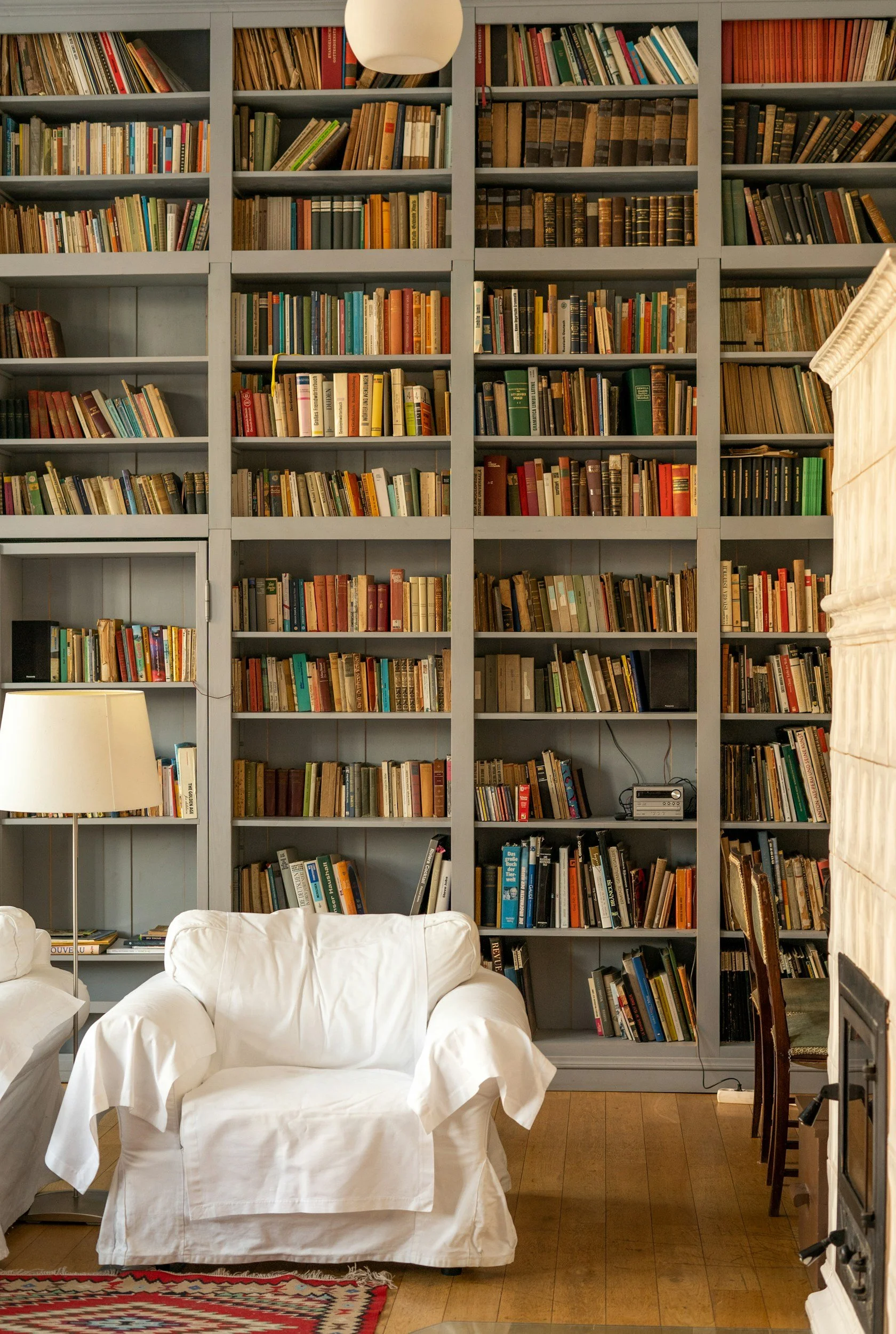 Living room with a large bookshelf filled with books, a white armchair, a table lamp, a cozy spot.