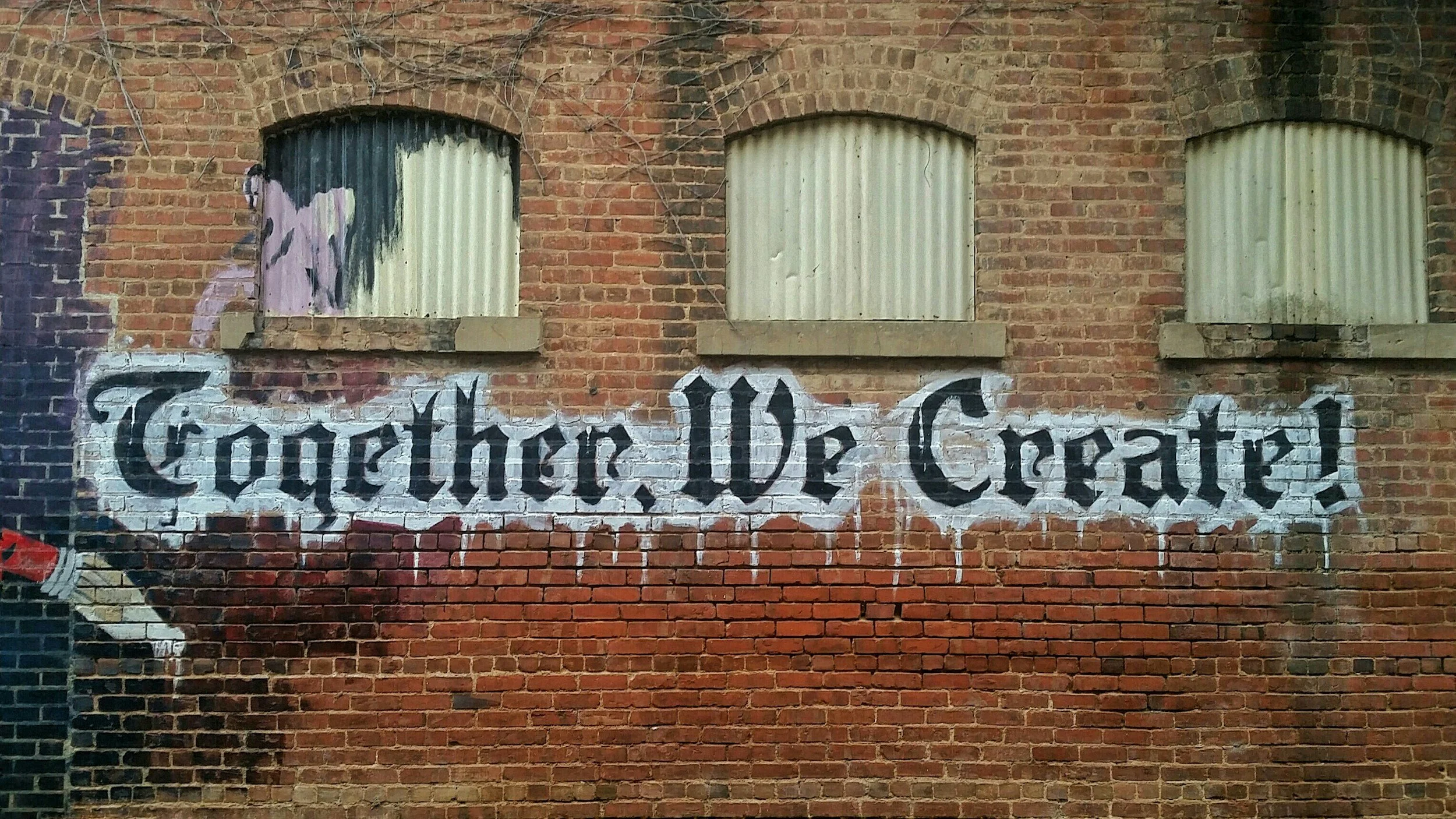 A brick wall with three boarded-up windows and graffiti that reads 'Together Be Great' in black cursive lettering.