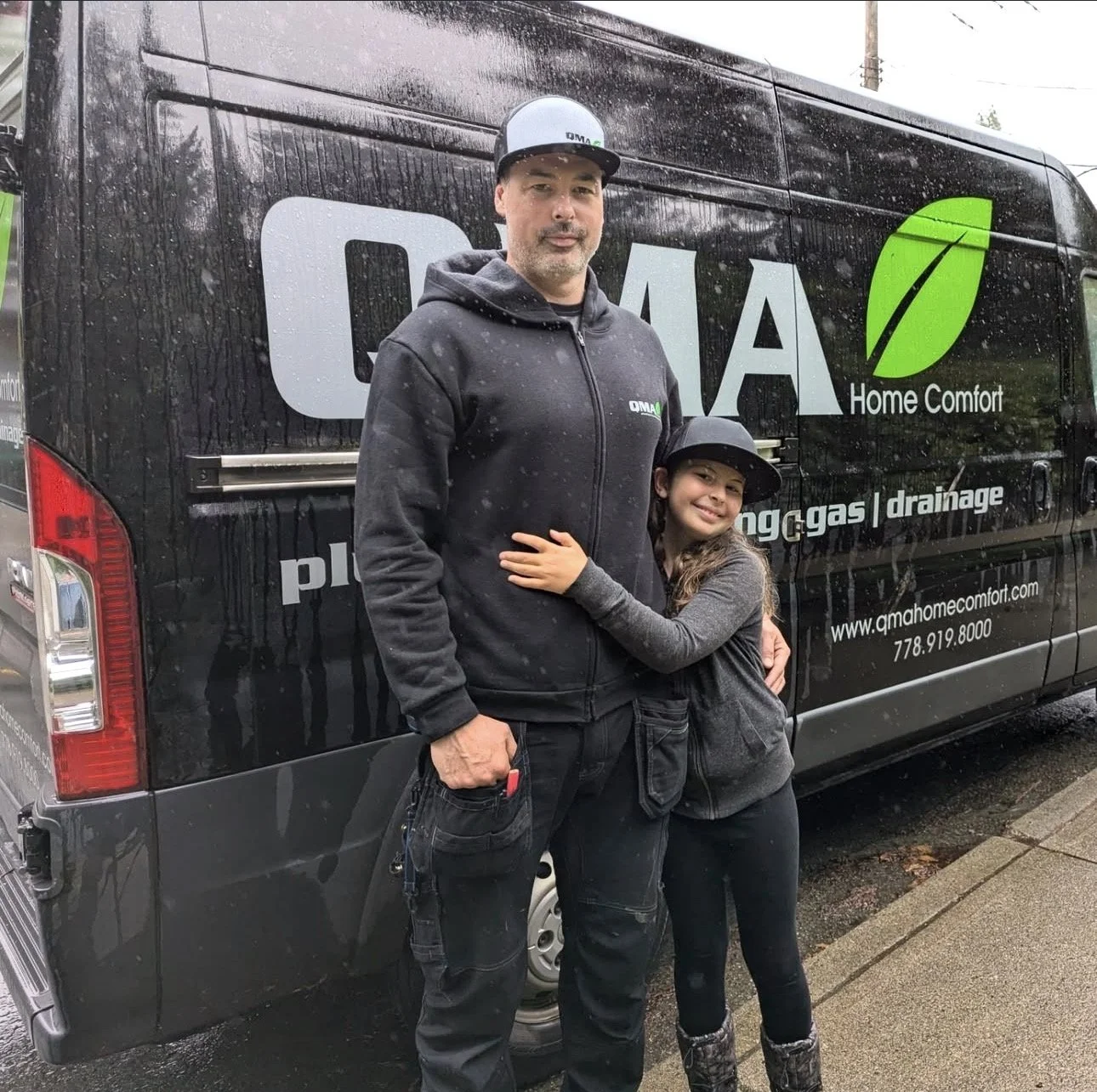 A man and young girl standing and hugging in front of a black company van with branding for QMA Home Comfort, specializing in natural gas and drainage services, on a rainy day.