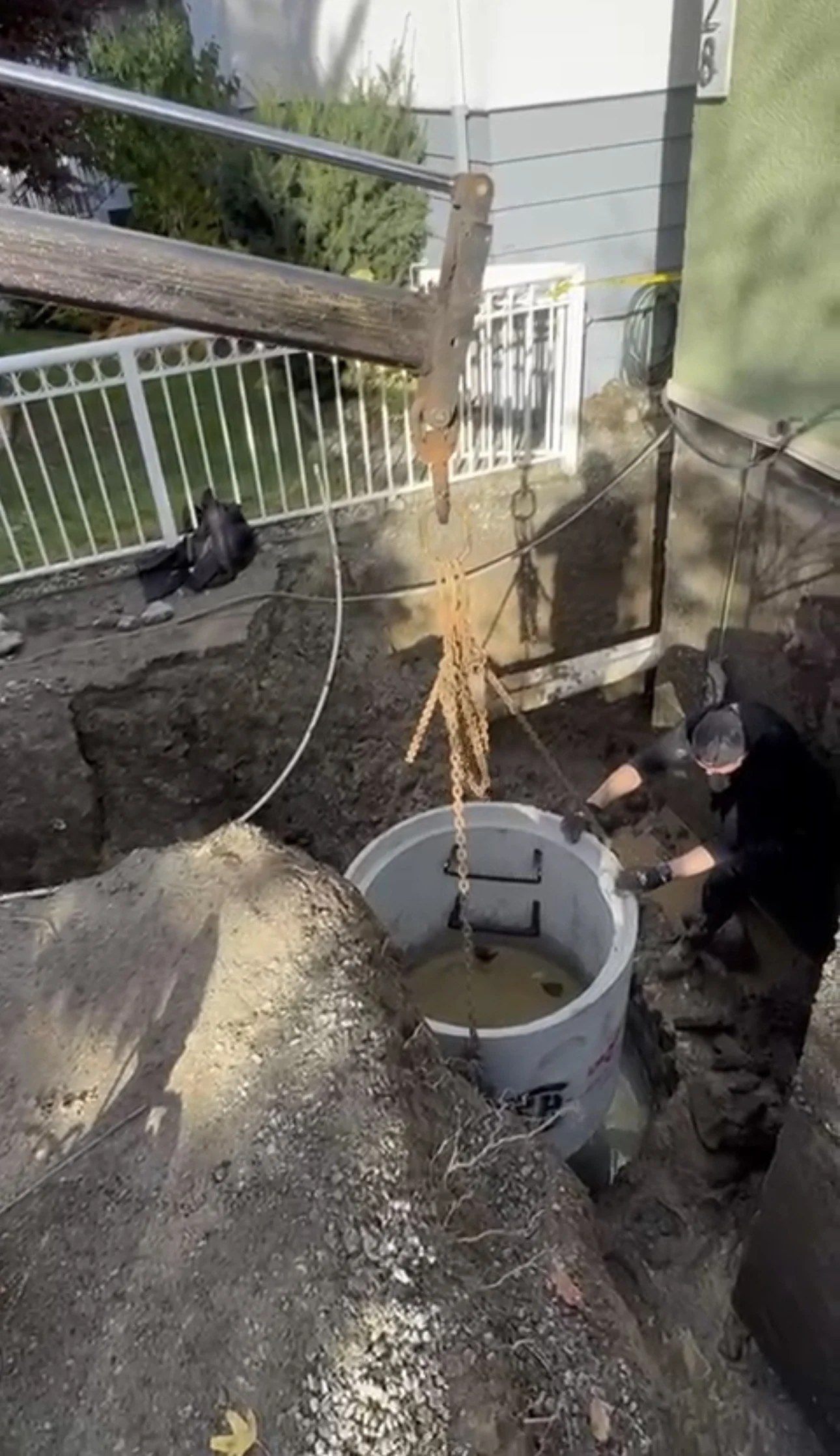 A construction worker is working in a trench, installing a large concrete pipe section. A chain hoist is lifting the pipe, and a black dog is sitting nearby on the surface above. There is dirt and excavation debris around the site, with a white fence and greenery in the background.