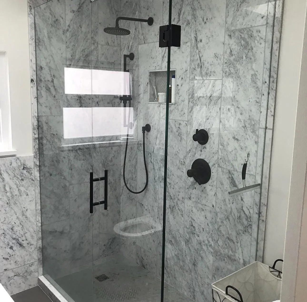 Modern shower with marble walls, black fixtures, a handheld showerhead and a rainfall showerhead, a built-in niche for toiletries, and a glass door, with a laundry basket outside.