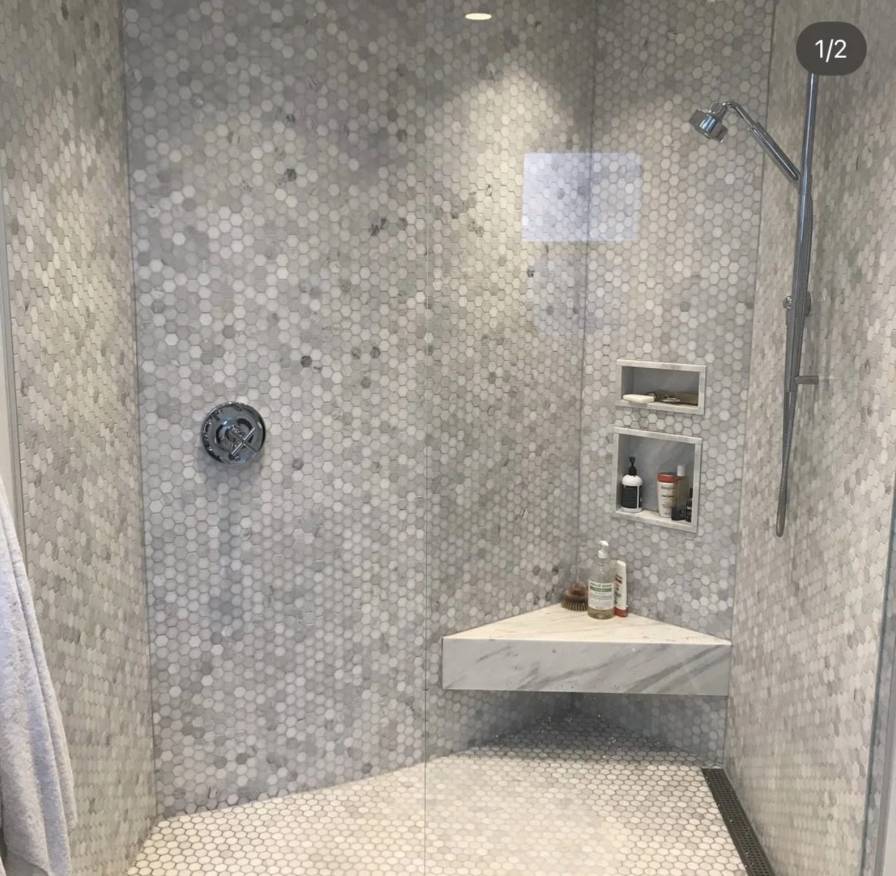 A modern walk-in shower with hexagonal white tile on the walls and floor, a built-in shelf with toiletries, a marble bench, and a rainfall showerhead.