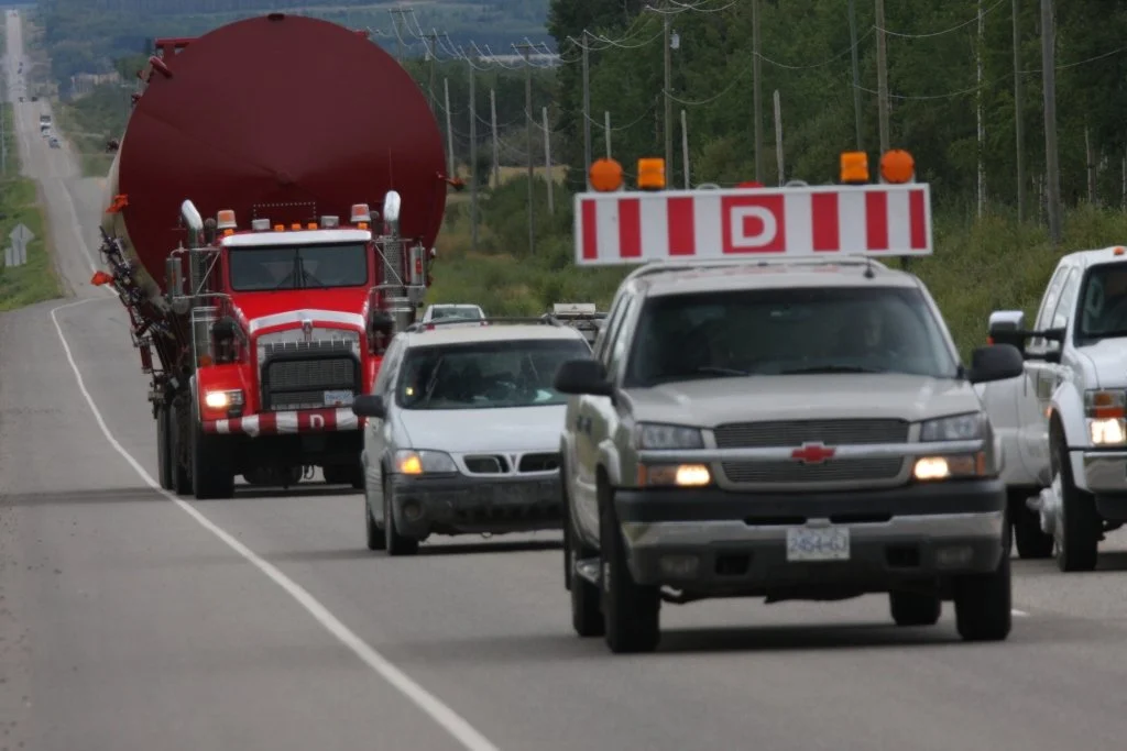 Line of vehicles including a large red tanker truck, a gray utility truck with a D sign, and several cars traveling on a rural highway surrounded by green trees and power lines.