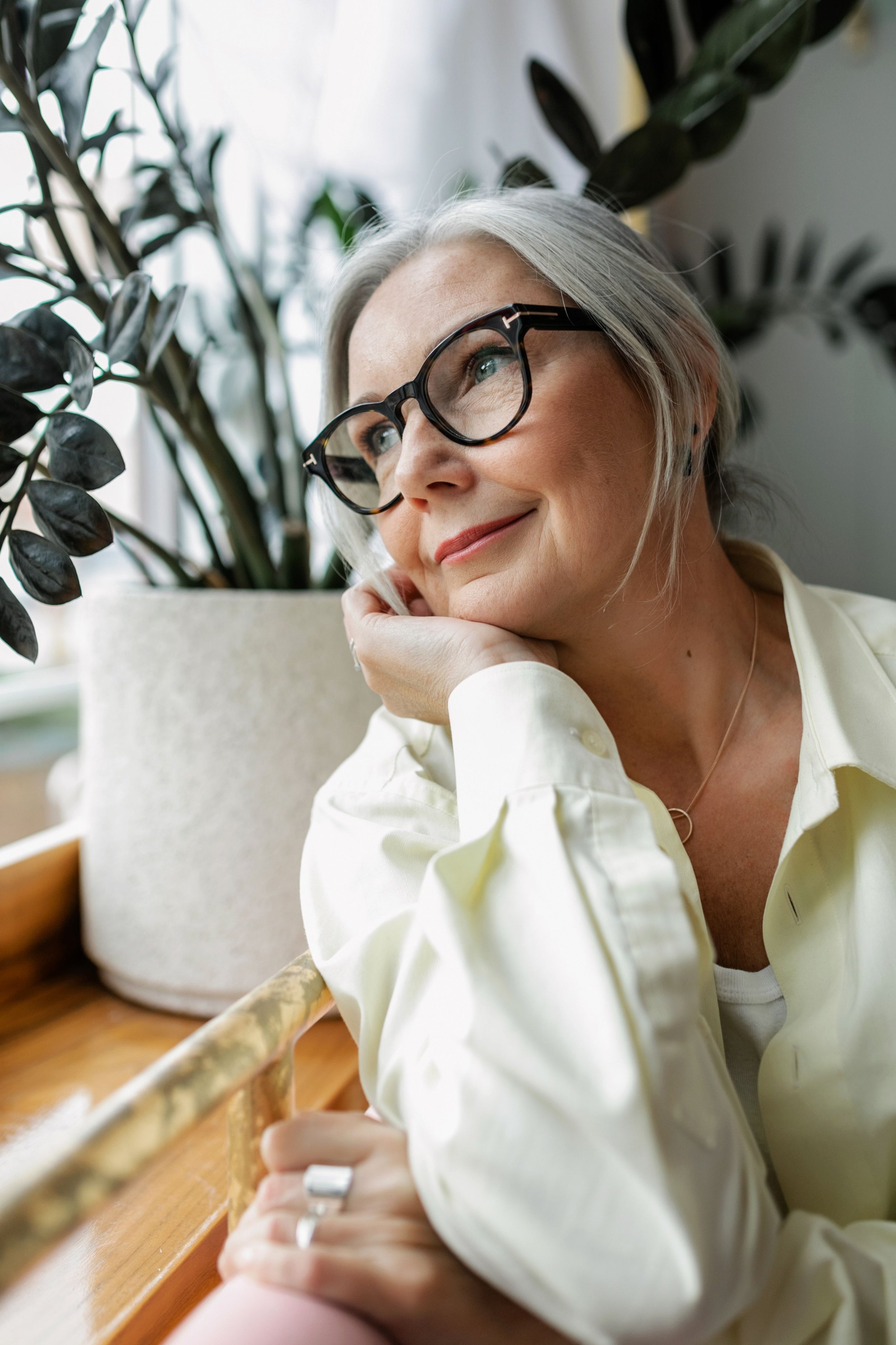 Mature woman with silver hair sitting by a window in natural light, showing natural mature skin.