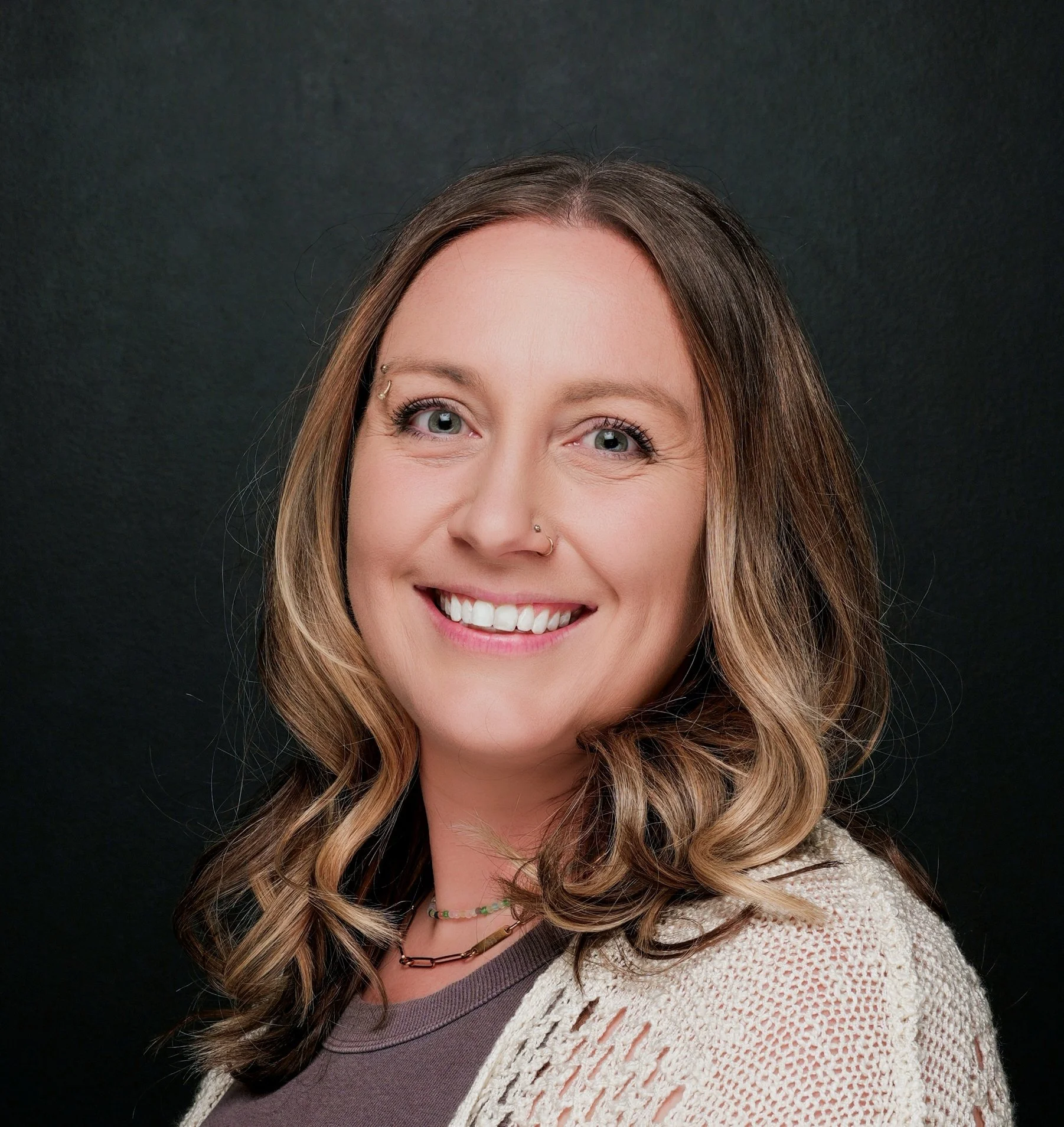 A smiling woman with wavy brown hair, wearing a gray top and a beige knit cardigan, against a black background.