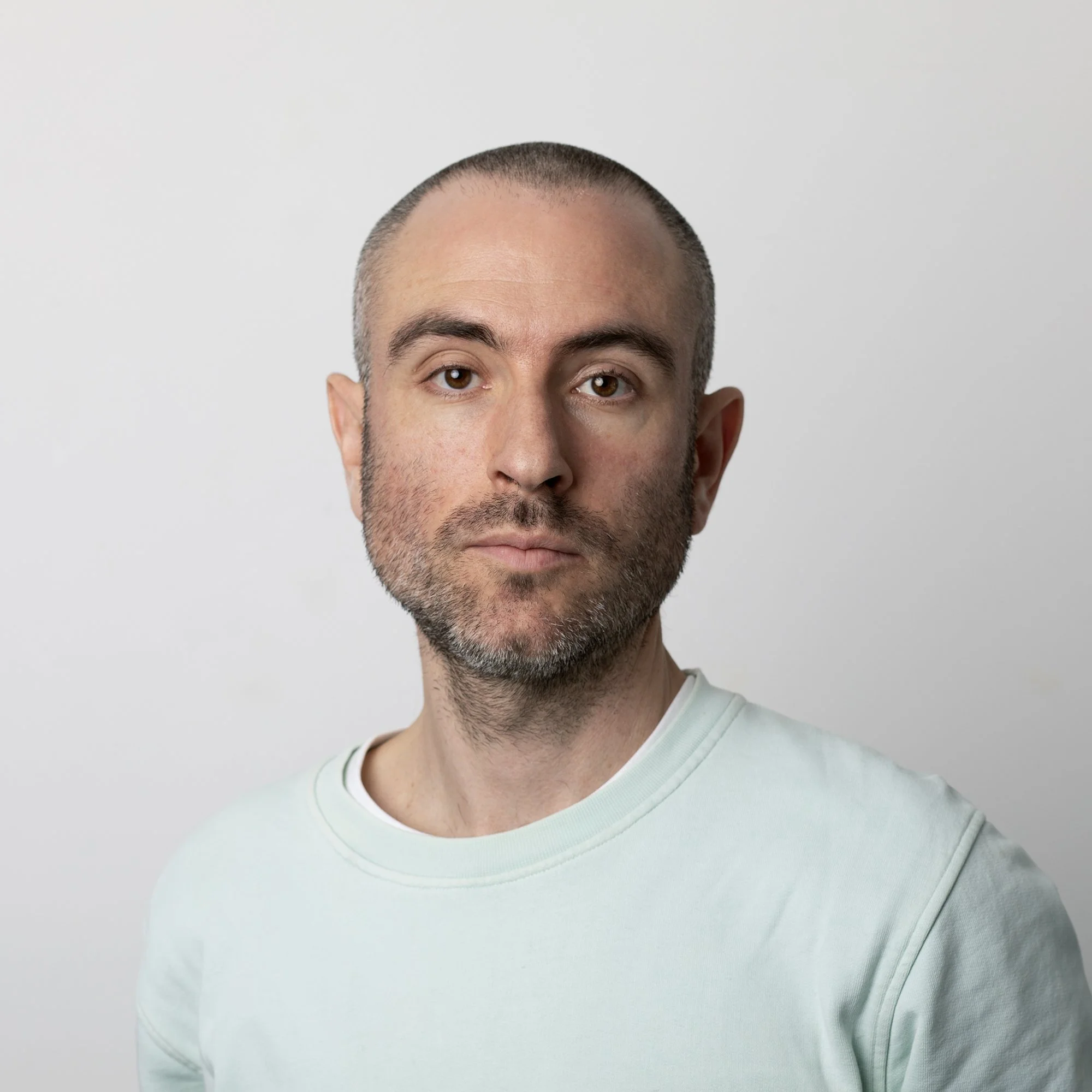 A man with a shaved head, beard, and mustache wearing a light-colored shirt against a plain white background.