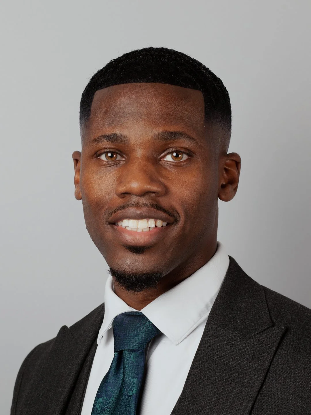 Professional headshot of a young man with short curly hair, wearing a dark suit, white shirt, and a blue tie, smiling in front of a plain light grey background.