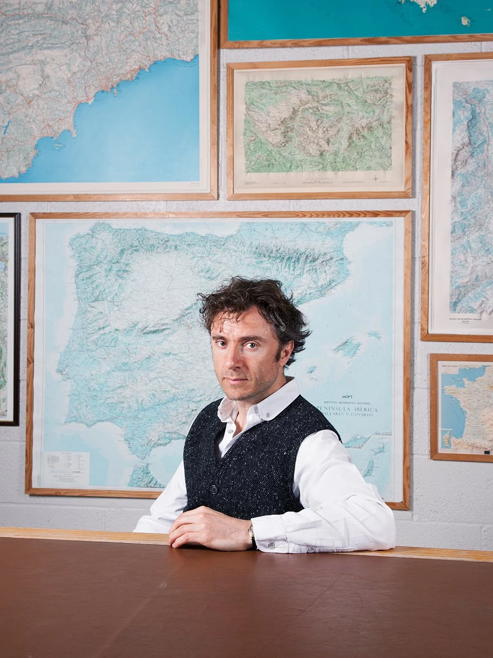 A man with dark, curly hair and a light complexion sitting at a table in front of a wall decorated with framed maps of various regions, mostly in blue and green colors.