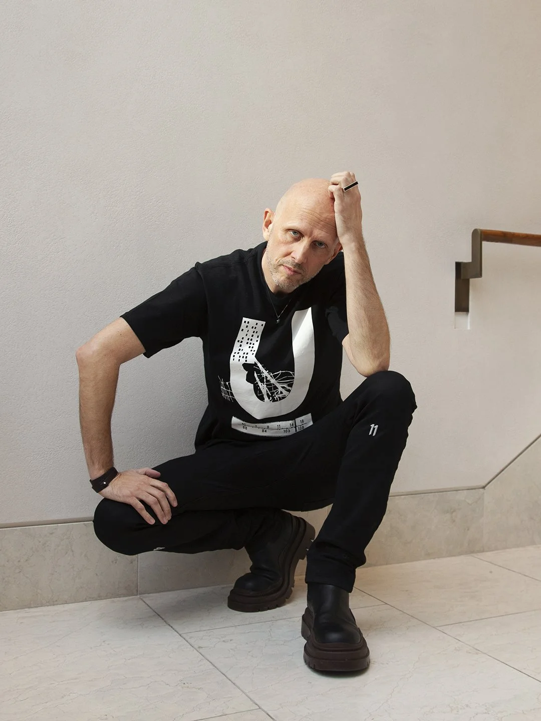 A man crouching down on the floor, resting his elbow on his knee and holding his head with his hand, looking directly at the camera. He has a bald head, light skin, and is wearing a black t-shirt with a graphic design, black pants, and black boots. The background features a plain, light-colored wall and a staircase railing.