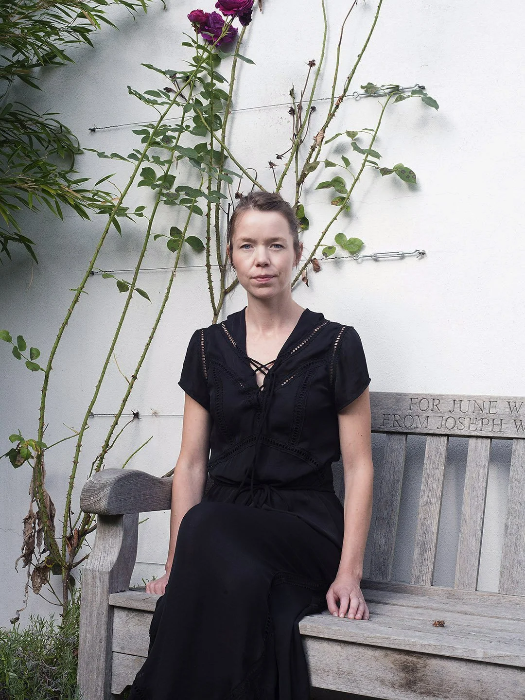 A woman in a black dress sitting on a wooden bench outdoors, with a white wall and flowering vines behind her.