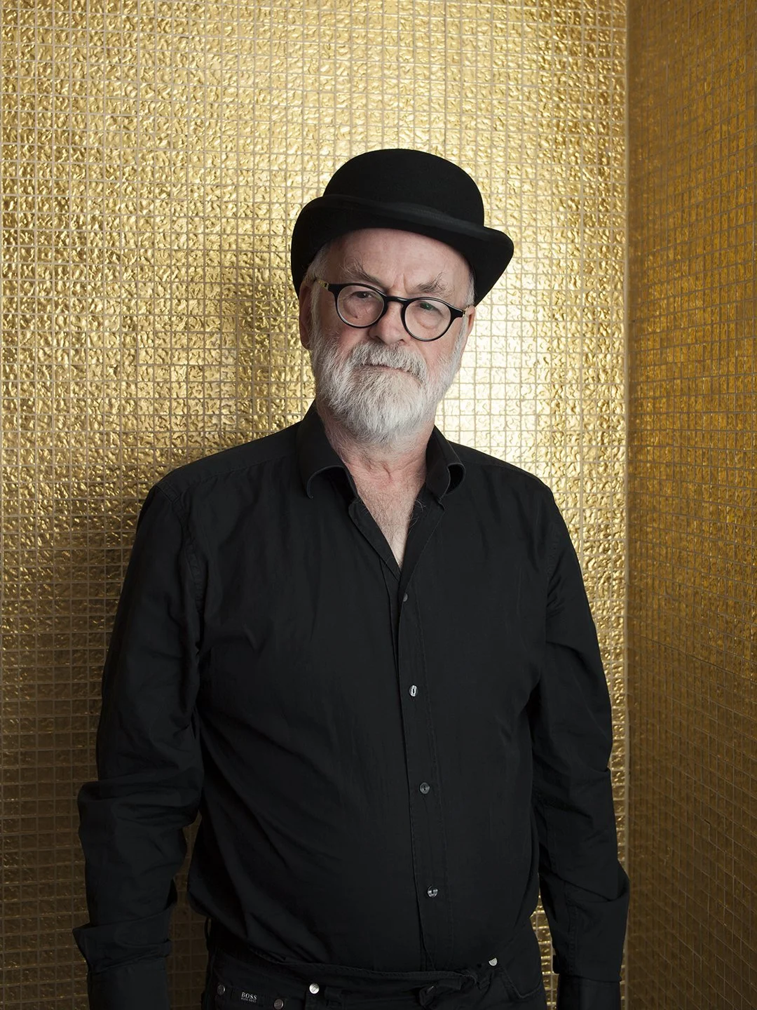 Older man with gray hair and a beard wearing black glasses, a black shirt, and a black hat standing in front of a gold textured wall.