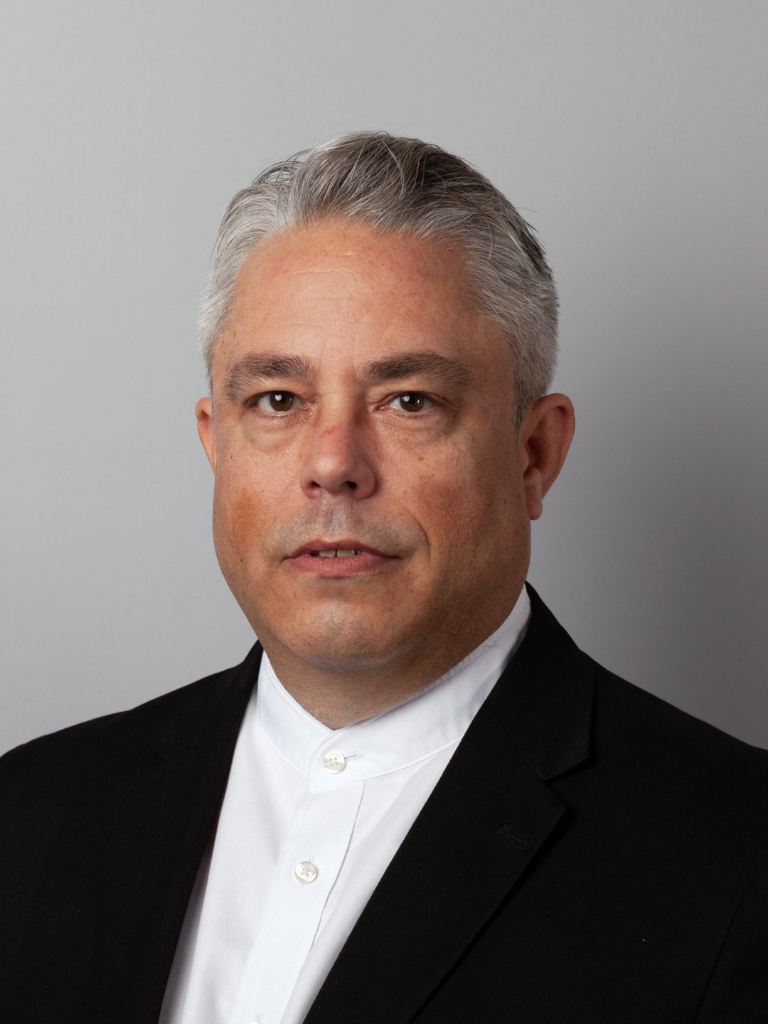 Professional portrait of a middle-aged man with gray hair, wearing a black suit and white shirt, against a gray background.
