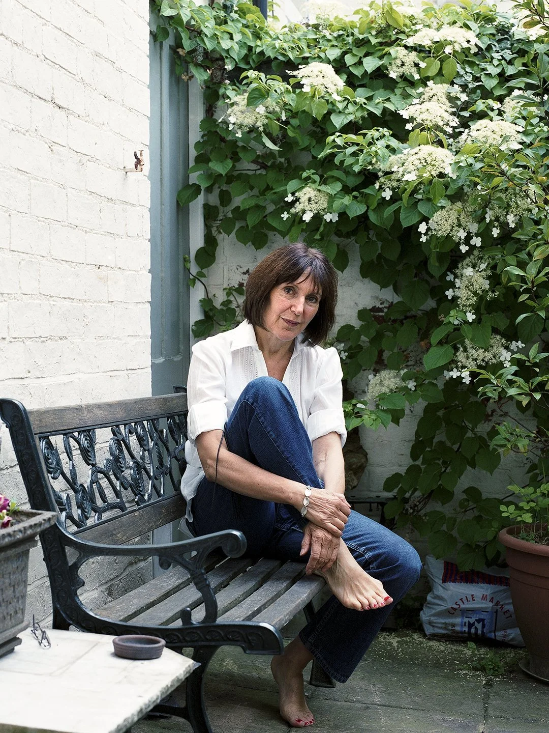 A woman sitting barefoot on a garden bench, with her arms wrapped around her legs, in a lush outdoor garden with green plants and white flowers.