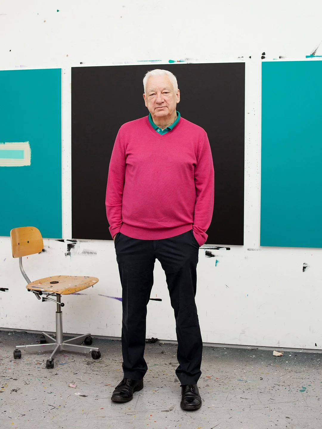 An elderly man standing with hands in pockets in front of an abstract art display, wearing a pink sweater over a teal polo shirt, black pants, and black shoes, in an art studio with a wooden chair and paint-splattered floor.