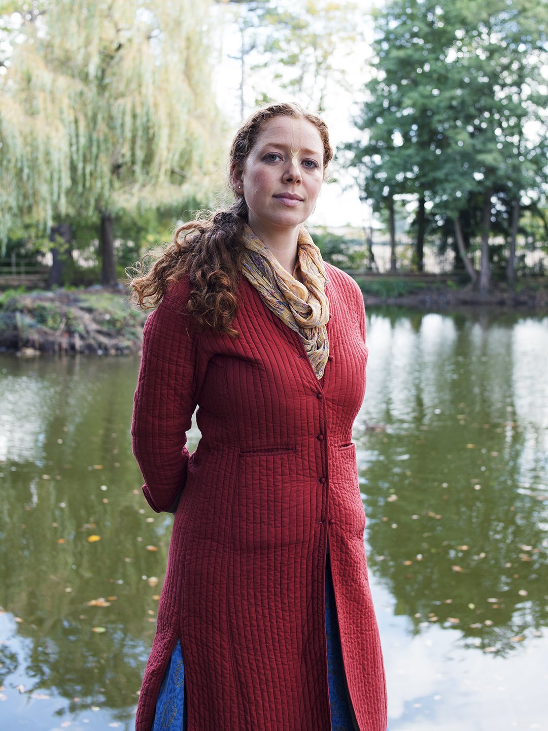 A woman with curly red hair stands outdoors with a pond and trees in the background, wearing a long red quilted coat and a patterned scarf.