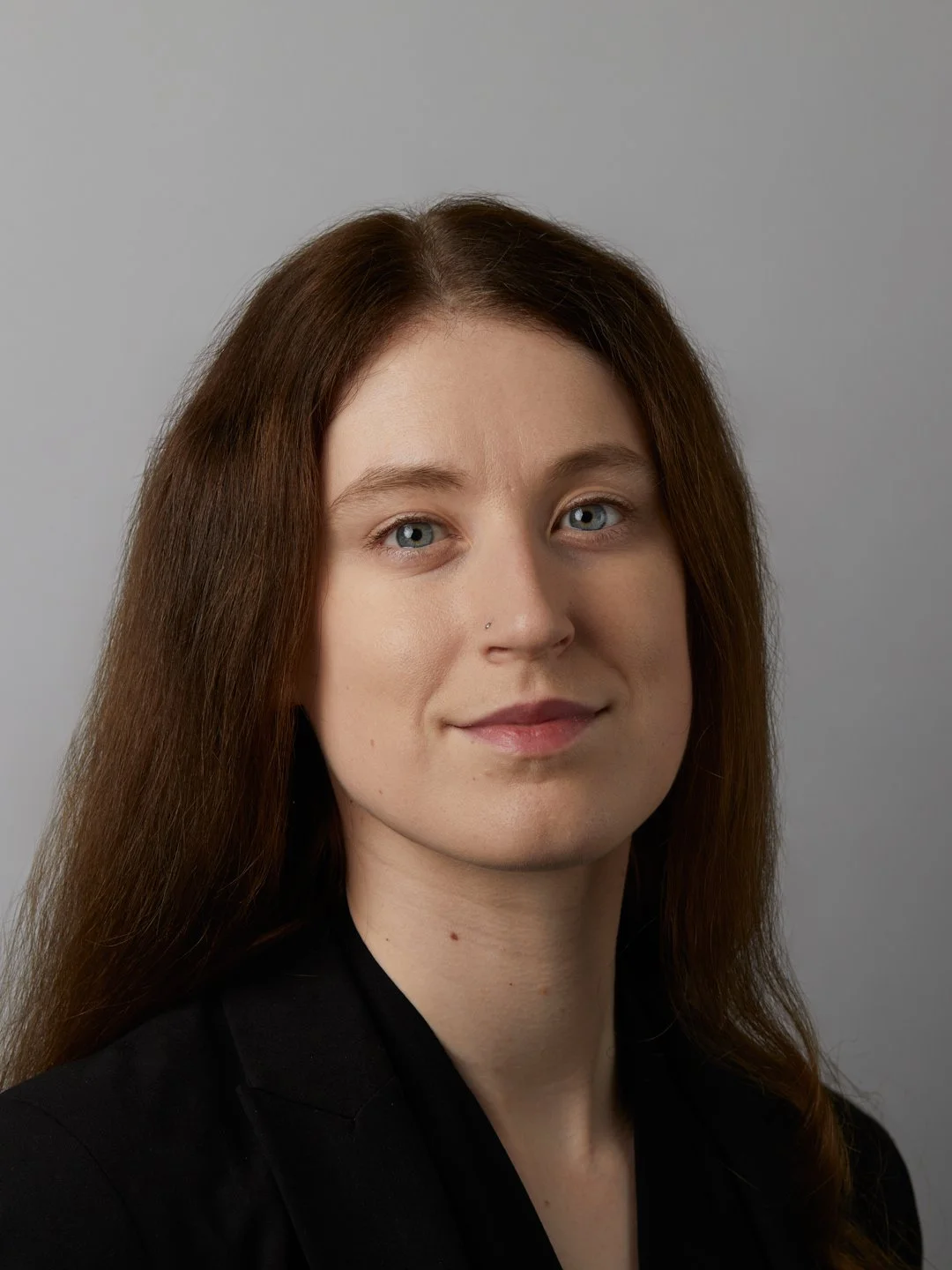 Portrait of a woman with long brown hair, blue eyes, wearing a black blazer, against a plain light gray background.