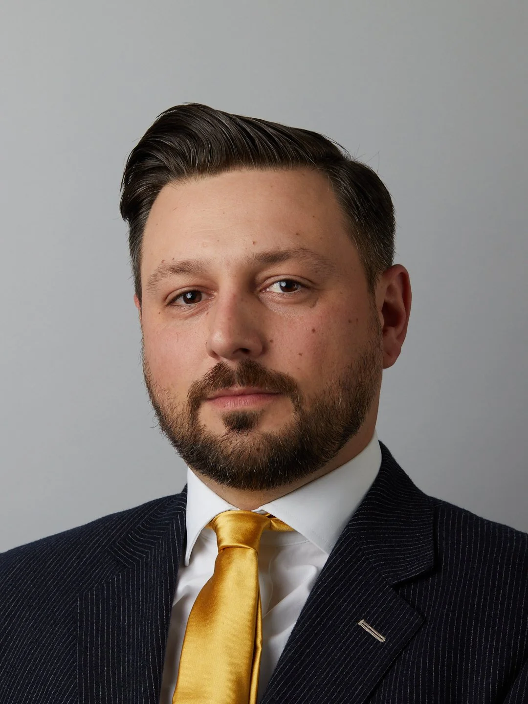 Portrait of a man with dark hair, beard, dressed in a business suit with a yellow tie, against a plain grey background.