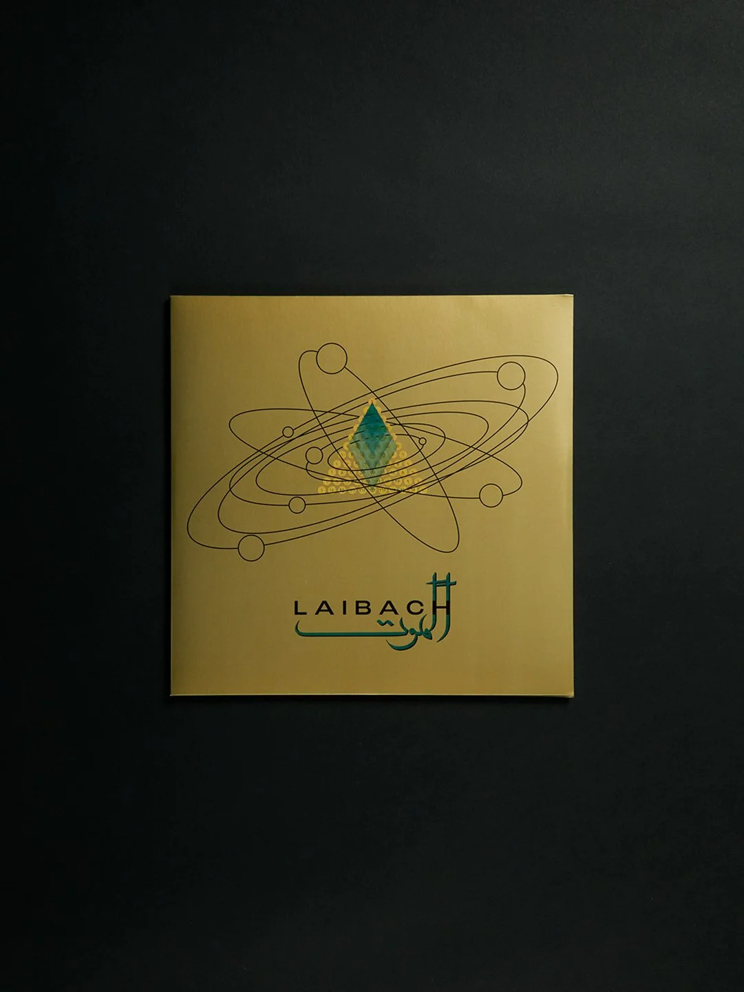 A yellow square paper with a scientific atom illustration and a gem-like crystal in the center. The word 'LAIBACH' is printed in black and there is Arabic text below it.