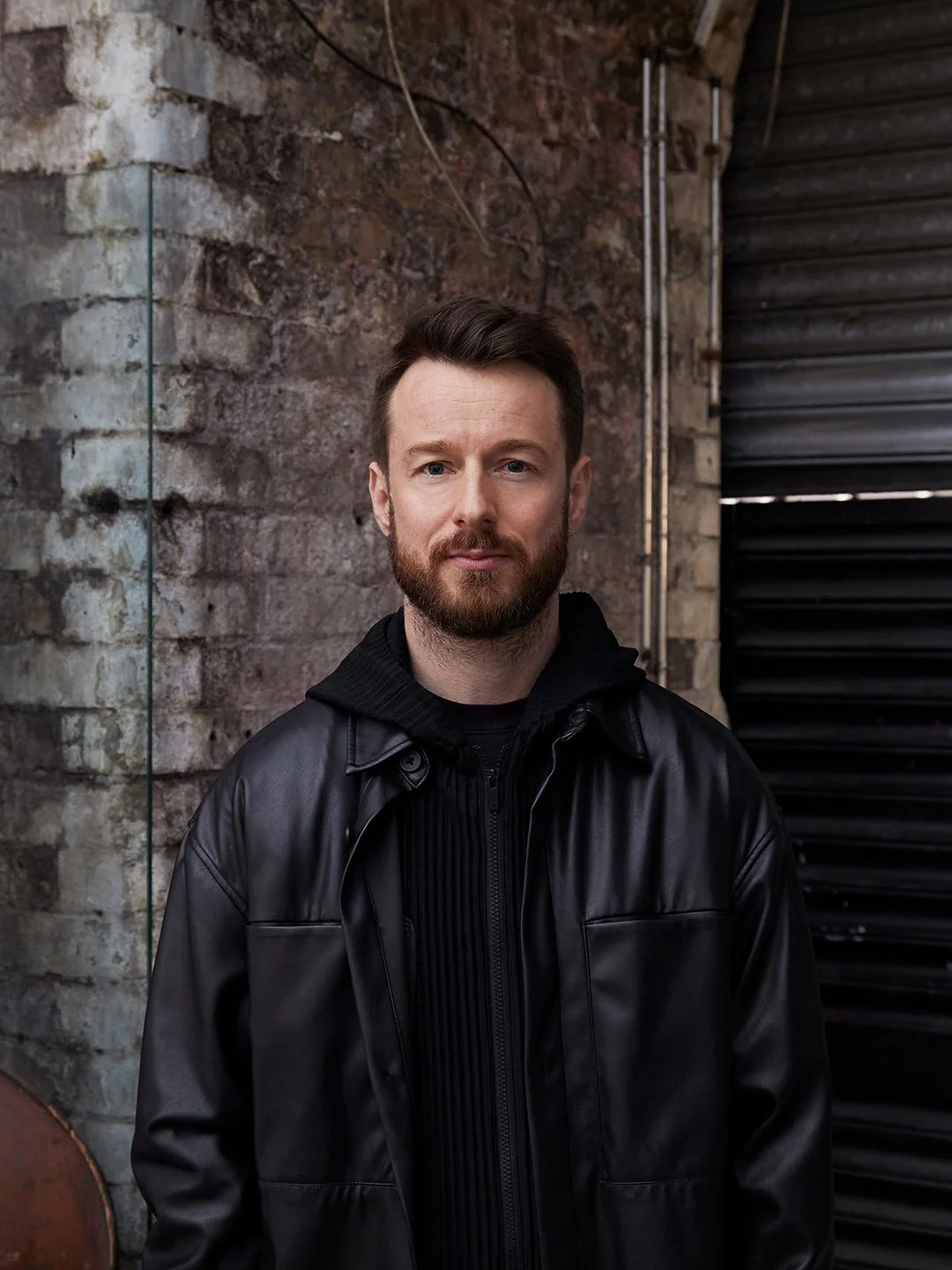 A man with a beard and short brown hair standing outdoors against a brick and metal wall, wearing a black leather jacket over a black hoodie.