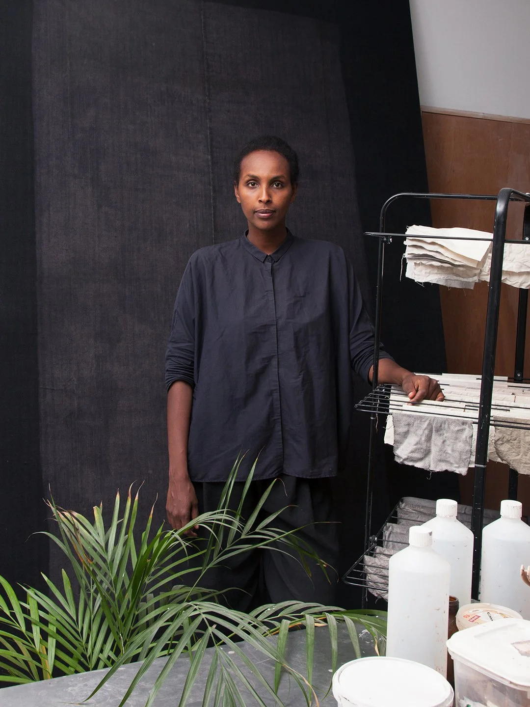 A woman standing with a serious expression in front of a dark background, wearing a dark blouse, with a plant in the foreground and a shelving unit with white bottles and fabric in the background.