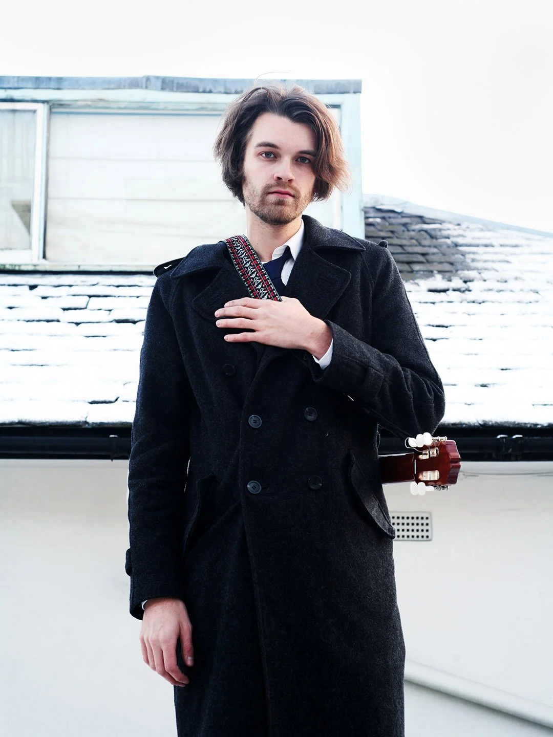 Young man with shoulder-length hair, wearing a black coat, standing outdoors in front of a snow-covered roof and cloudy sky, holding a guitar strap.