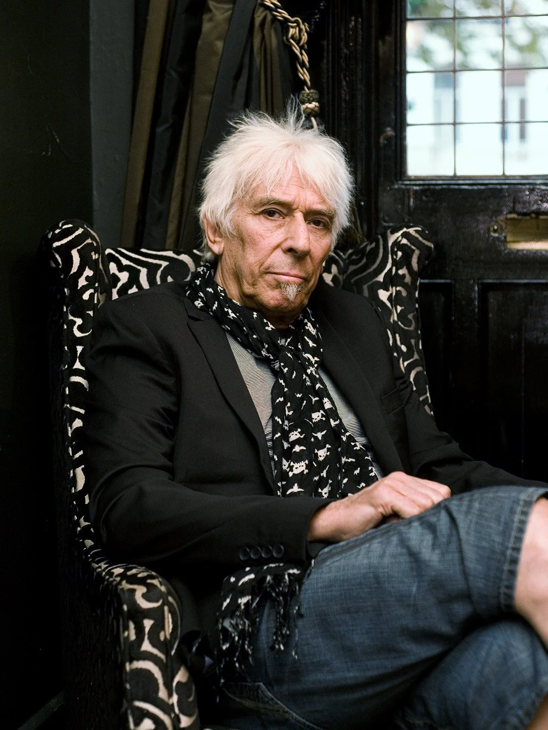 An older man with white hair and a beard, wearing a black blazer, patterned scarf, and jeans, sitting in a black and white patterned armchair in a dark room near a window.