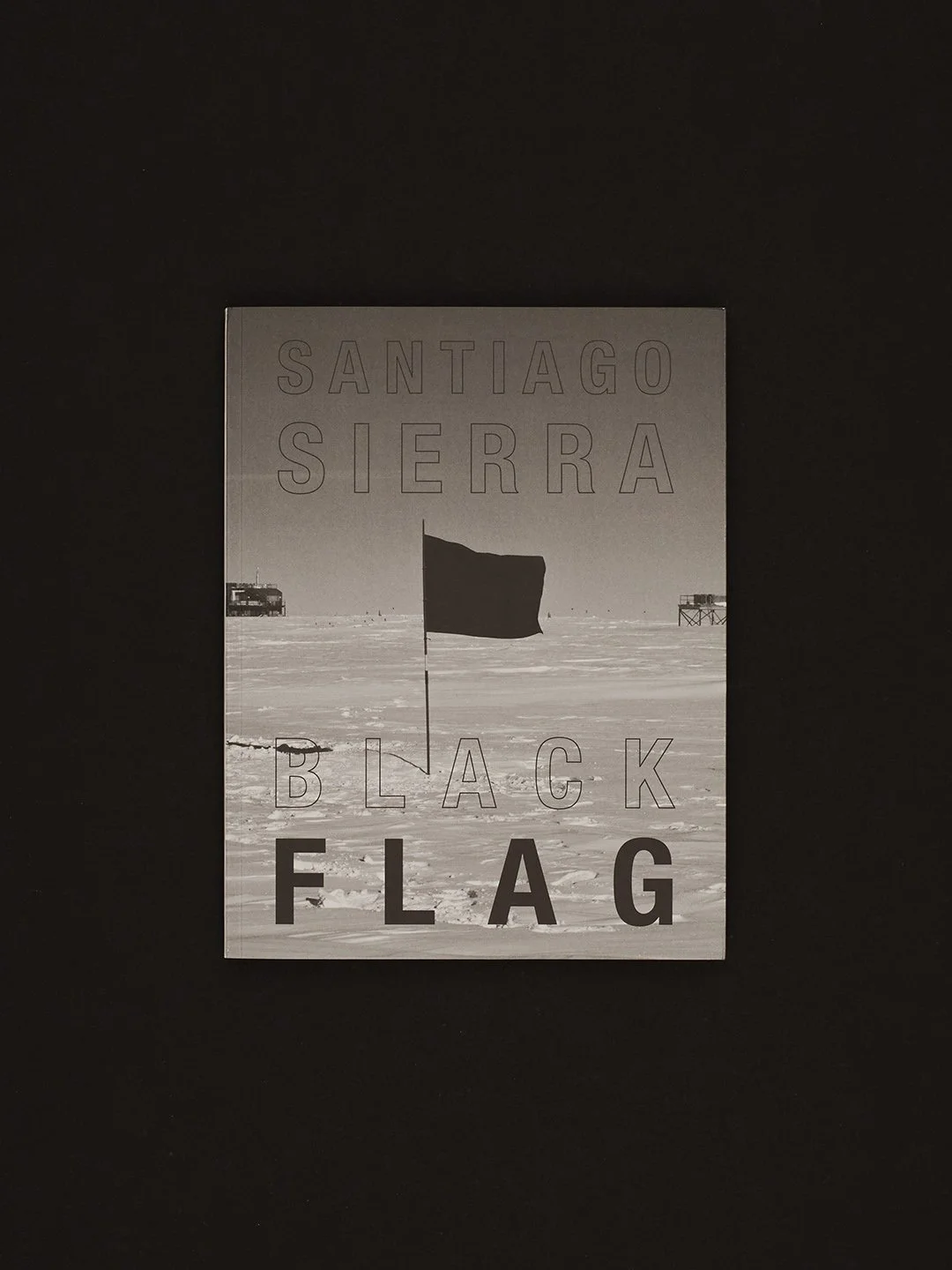 Book cover titled "Santiago Sierra Black Flag" with a desert scene, a black flag on a pole, and structures in the background, photo in black and white.