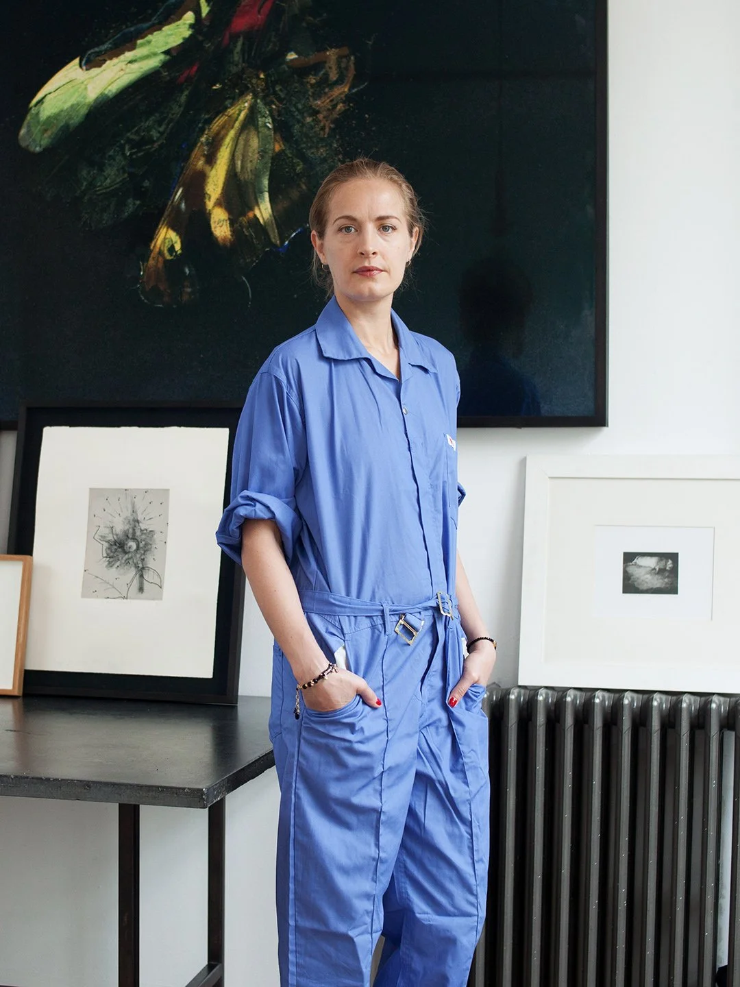 A woman in a blue jumpsuit standing in an art gallery with framed artwork and a large abstract painting behind her.