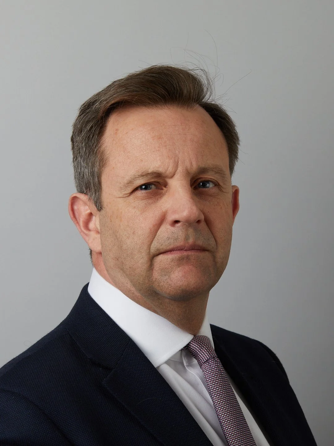 Close-up portrait of a middle-aged man in a suit, with short brown hair, blue eyes, and a serious expression, against a plain gray background.