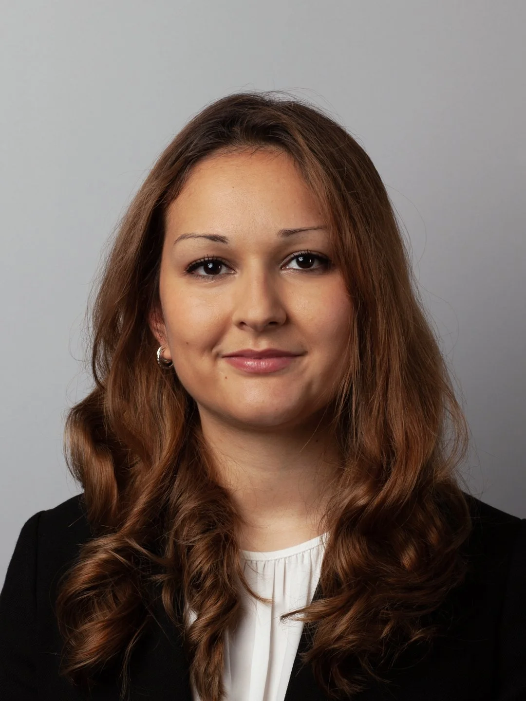 A young woman with long, wavy brown hair, wearing a black blazer and white top, smiling softly at the camera against a plain gray background.