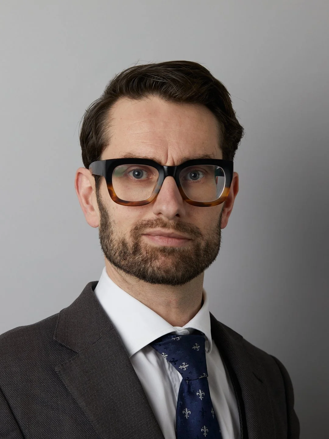 A man with dark hair, beard, and glasses wearing a suit with a tie, looking serious at the camera against a plain gray background.