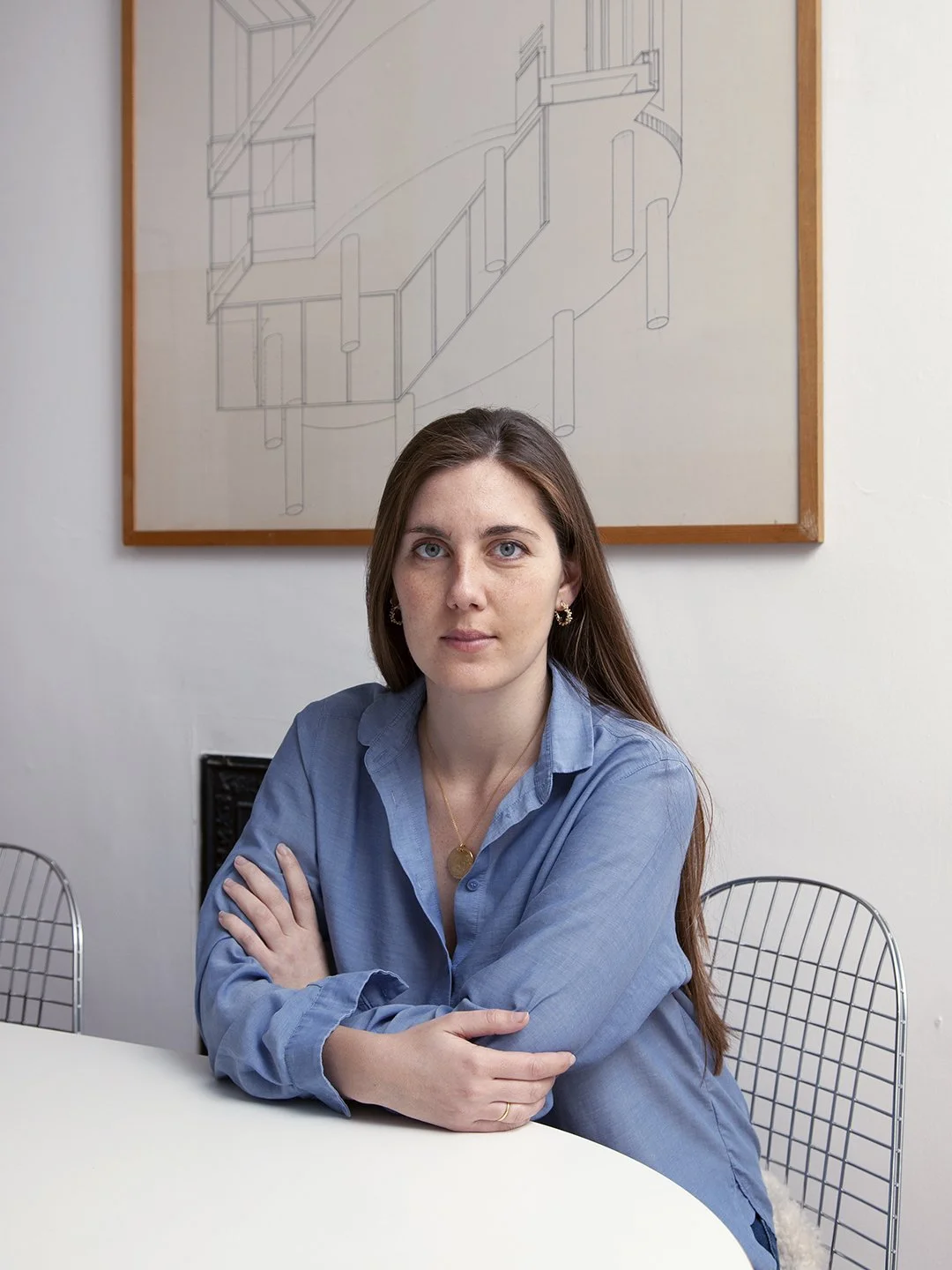 A young woman with long brown hair, wearing a blue button-up shirt, sitting at a white table with her arms crossed. There is a framed architectural drawing on the wall behind her, and a wire chair is partially visible to her left.