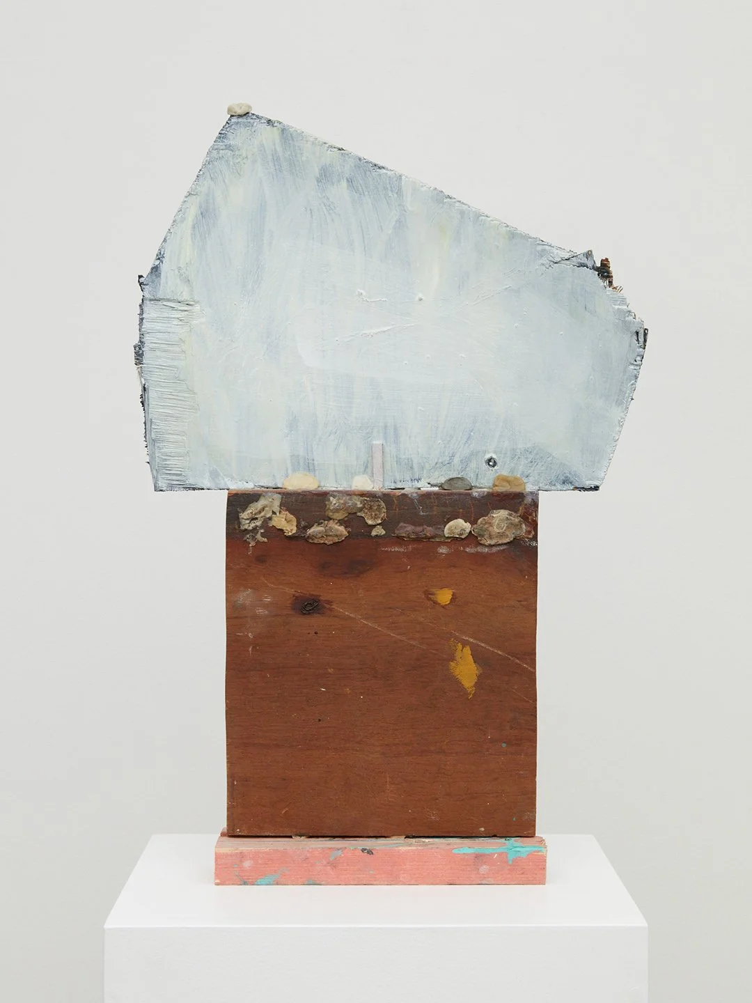 Abstract mixed media sculpture with a painted, textured upper section and a wooden lower section on a white pedestal.