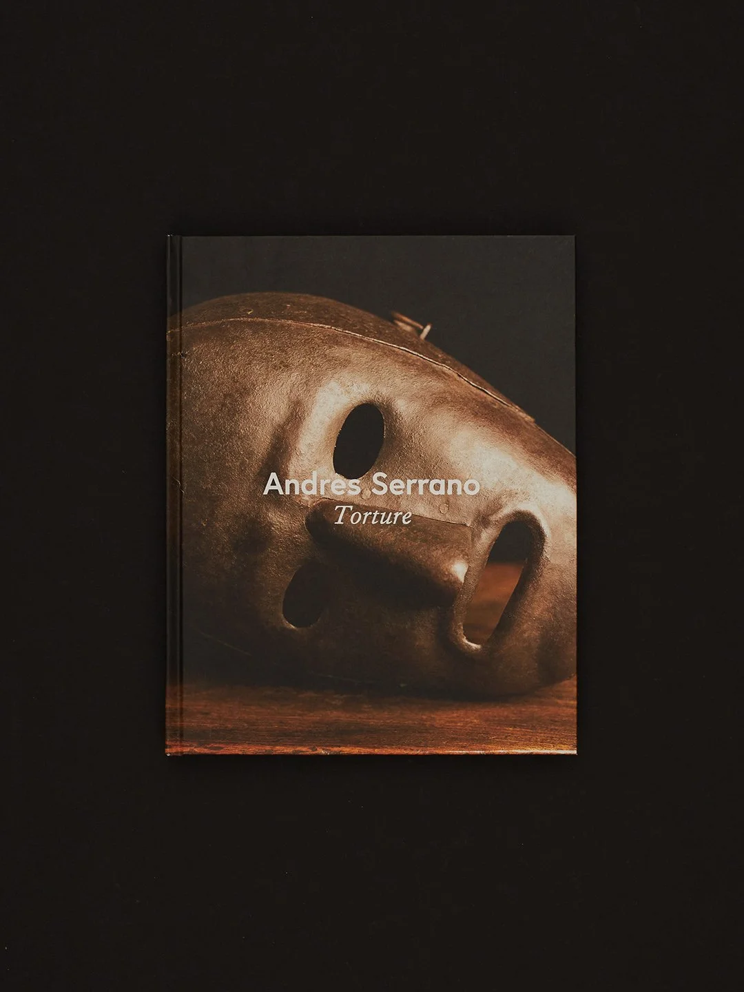 A book titled 'Torture' by Andres Serrano features a close-up photograph of a metallic mask with hollow eyes and a solemn expression, placed on a dark background.