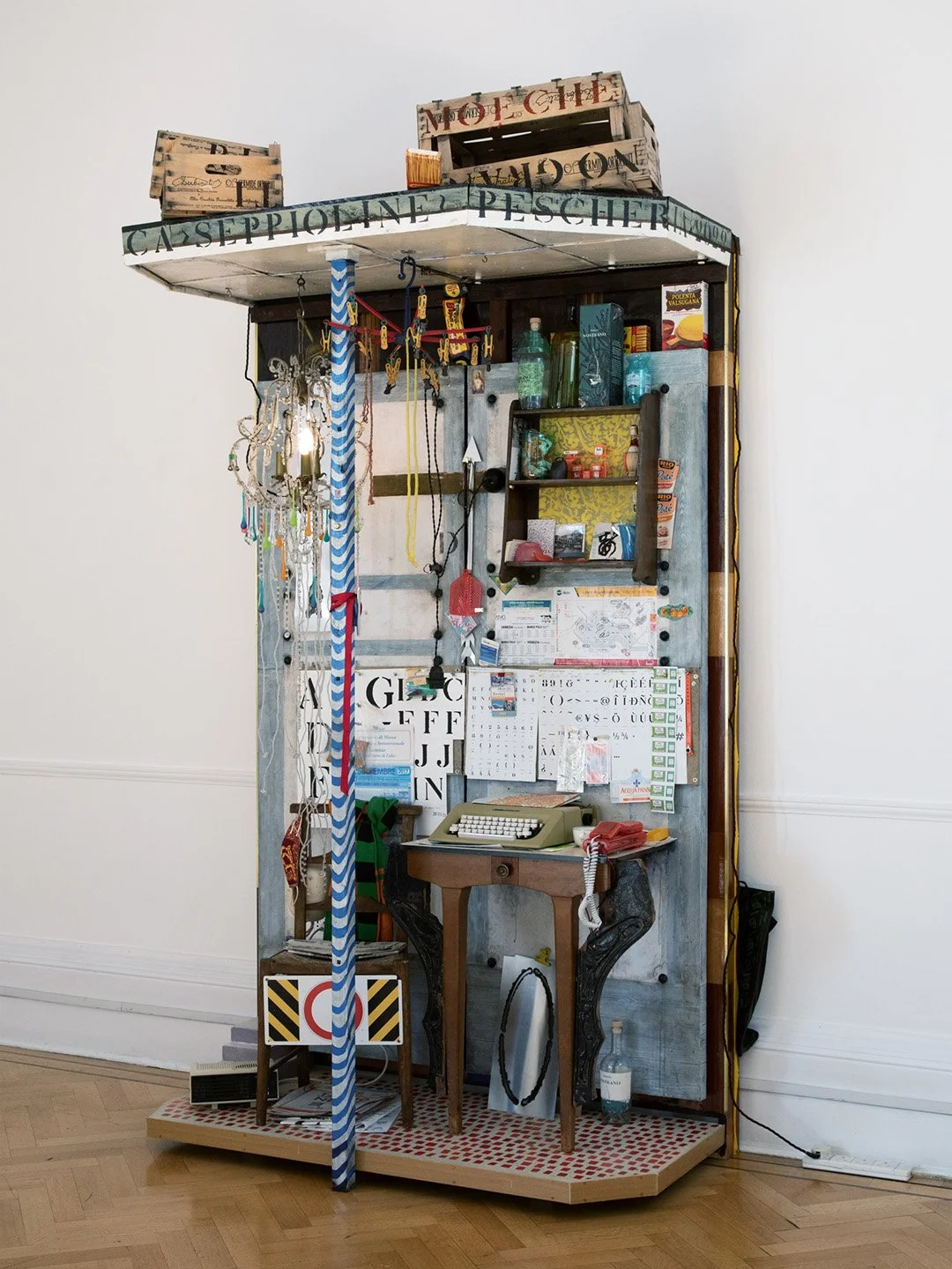 Eclectic art installation with vintage typewriter, cardboard boxes labeled 'MODICHE' and 'CASELPIOLINE PESCATORE,' a small shelf with bottles and decor, hanging jewelry, a colorful striped pole, and various papers and objects, all mounted on a platform in a room with white walls and wooden floor.