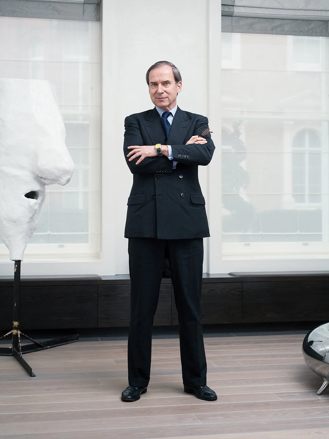 Man in a dark suit with a blue tie standing with arms crossed in a modern office, with abstract art sculpture to his left and large windows in the background.