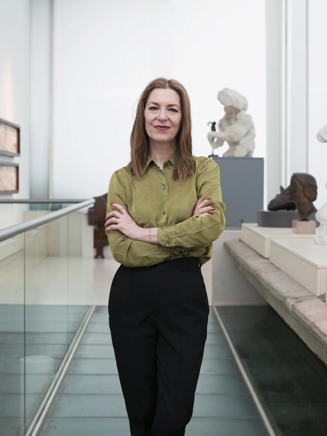 A woman with shoulder-length brown hair wearing a green button-up shirt and black pants standing in an art museum with sculptures and framed artworks in the background.