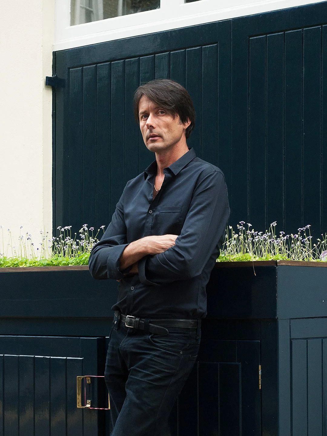 A man with dark hair wearing a black button-up shirt and black pants, standing outdoors with arms crossed against a dark blue fence and a flower bed with small purple flowers.