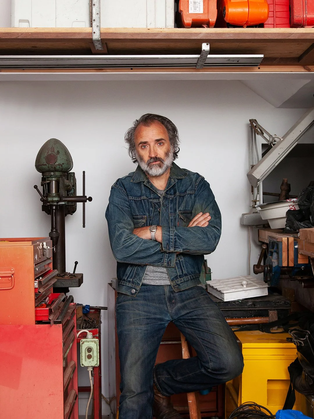 A middle-aged man with gray hair and a beard, wearing a denim jacket and jeans, standing in a workshop with arms crossed. The workshop has various tools and equipment, including a green drill press, orange toolboxes, and a yellow storage container.