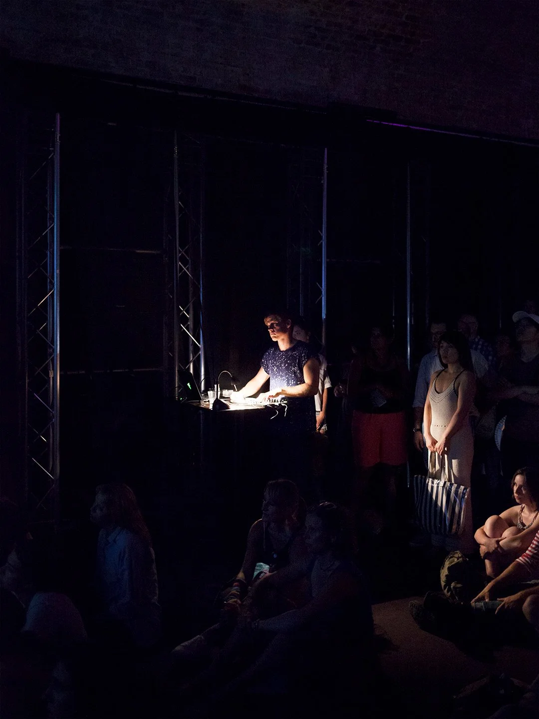 A DJ performs on stage with a laptop and music equipment while an audience watches in a dark, dimly-lit concert or event space.