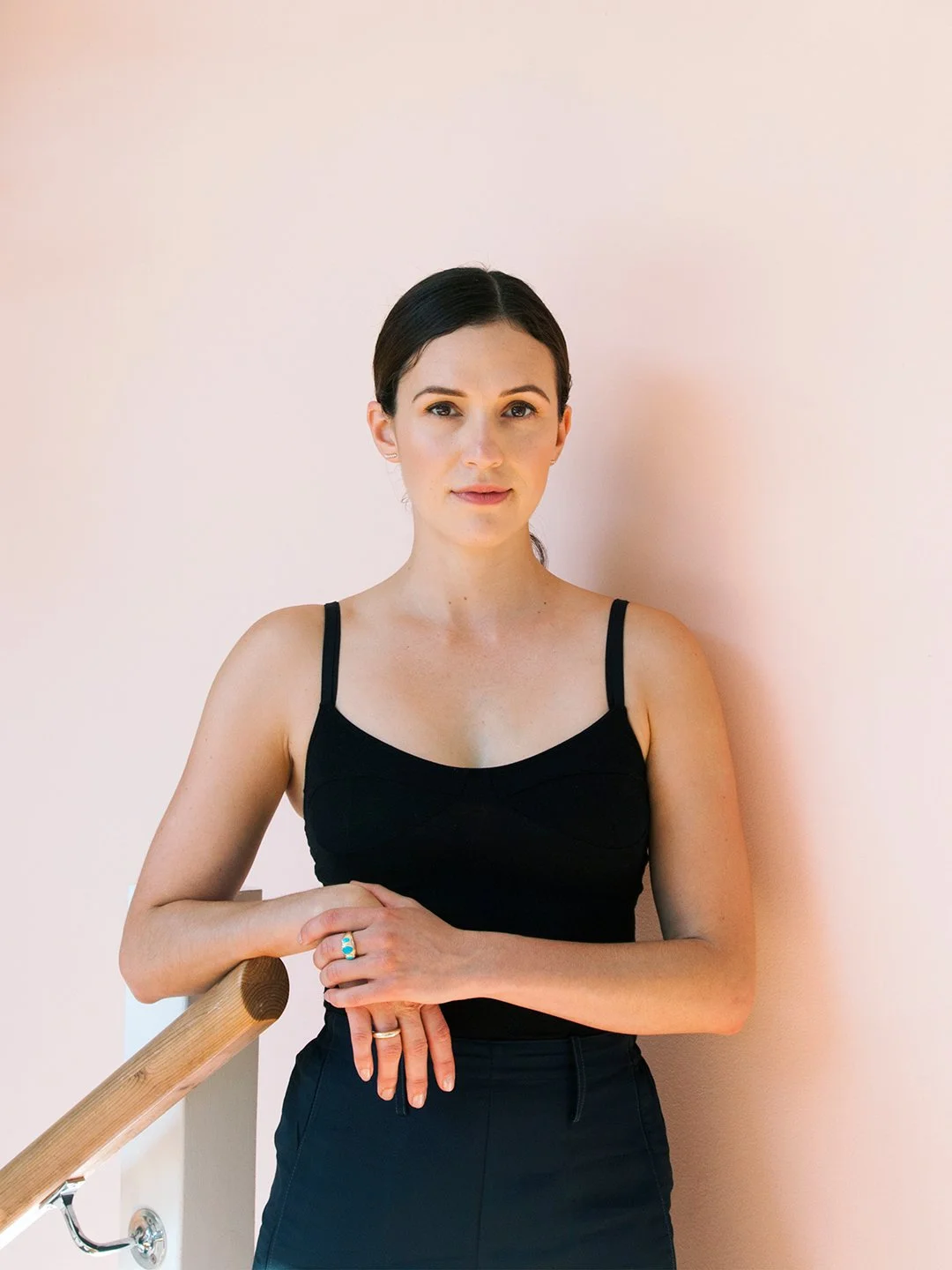 A woman with dark hair, wearing a black tank top and black pants, standing against a light pink wall, resting one hand on a wooden railing.