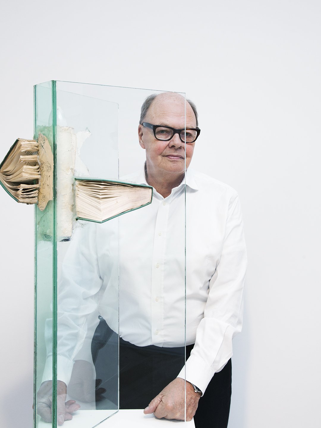 A man with glasses in a white shirt standing behind a glass display case containing old books. The case has glass panels on three sides and is positioned against a plain white wall.