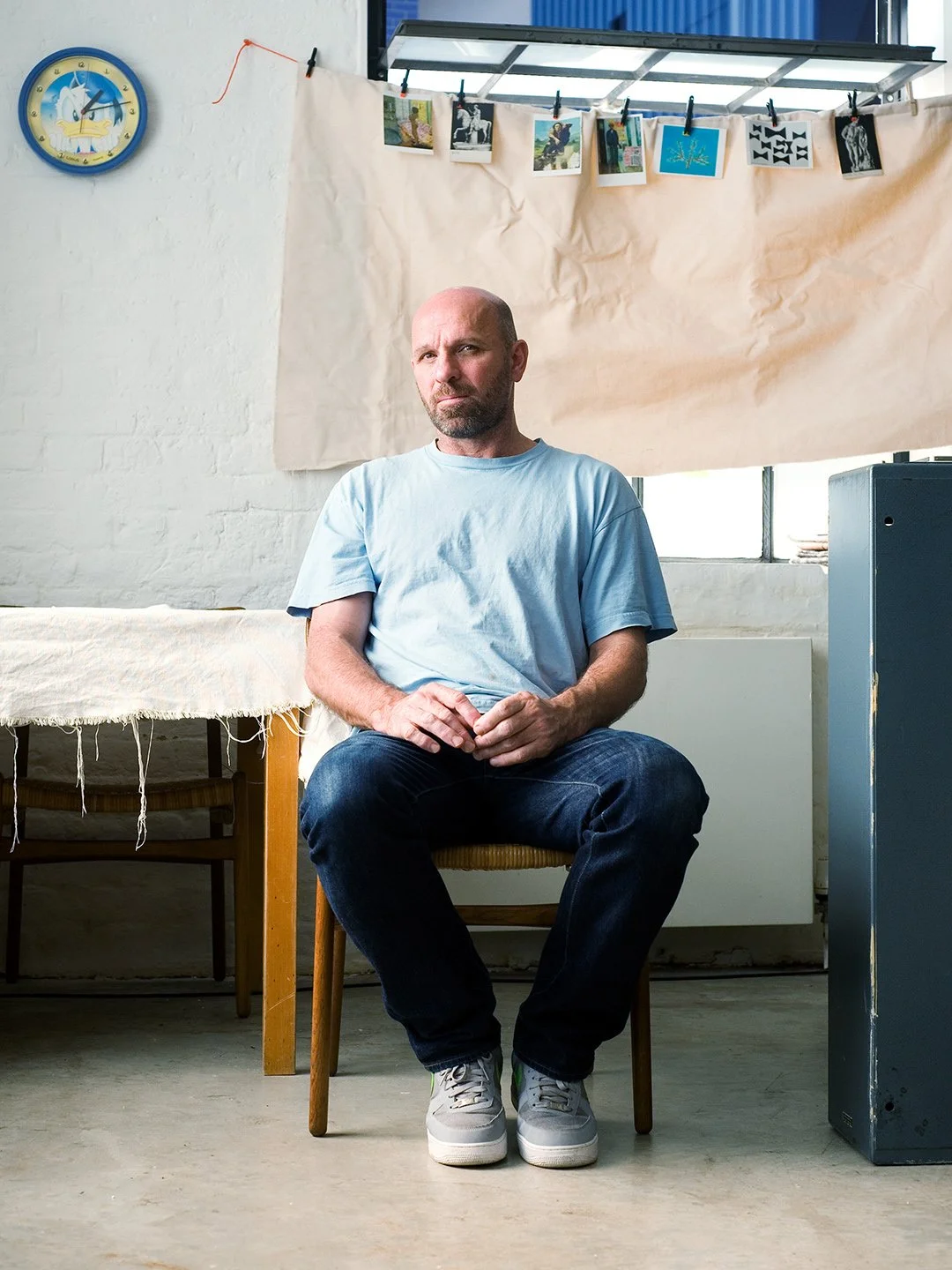 A man with a beard and bald head wearing a light blue T-shirt and jeans, sitting on a wooden chair with his hands clasped, in an indoor space with a white brick wall, a clock, and photographs hanging on a line behind him.