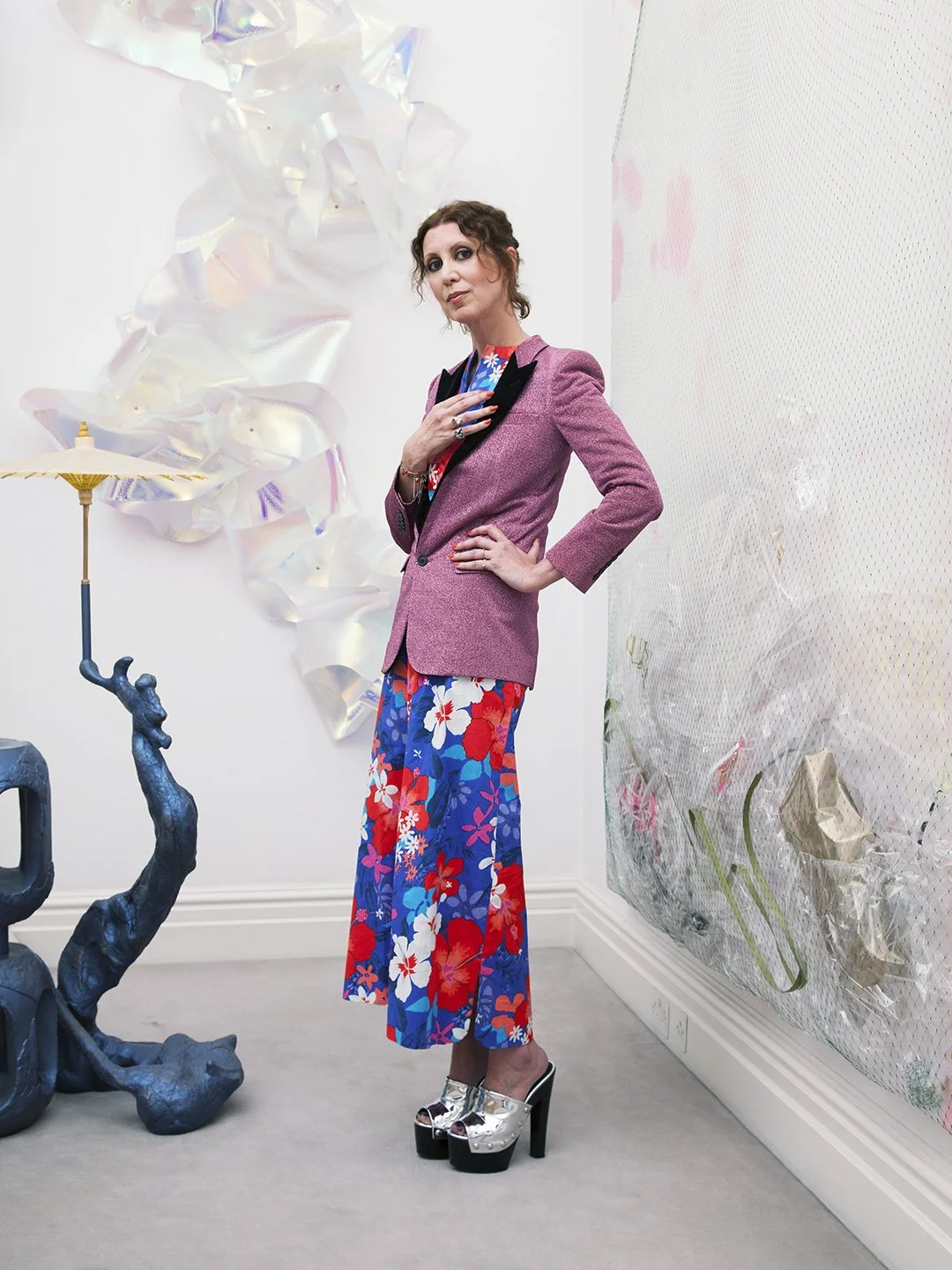 Woman in colorful floral pants and pink blazer standing indoors next to abstract art installations, wearing platform shoes.