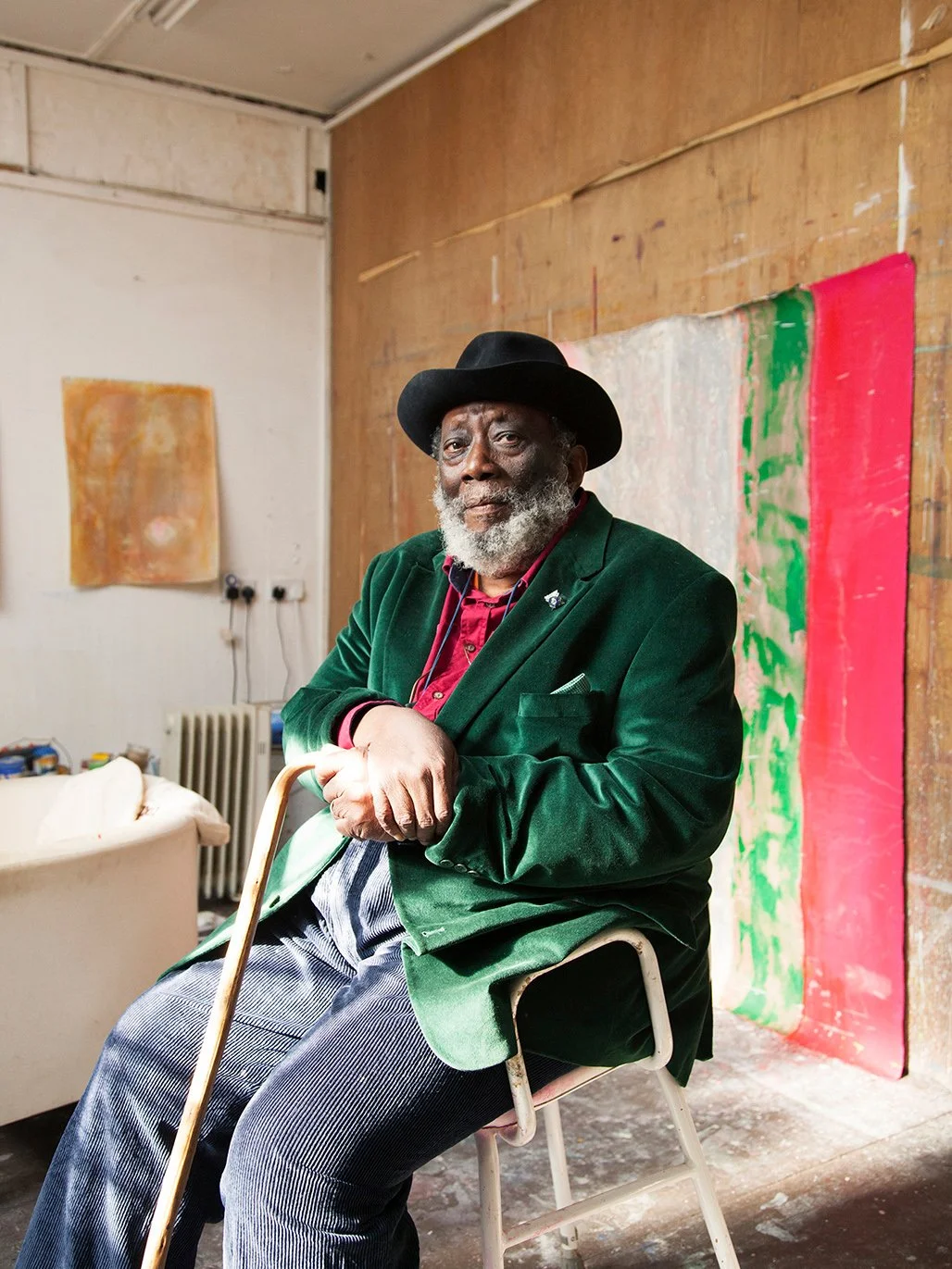 An elderly man with a gray beard, dressed in a green velvet blazer, red shirt, and striped pants, sitting on a chair in an art studio with abstract paintings in the background.