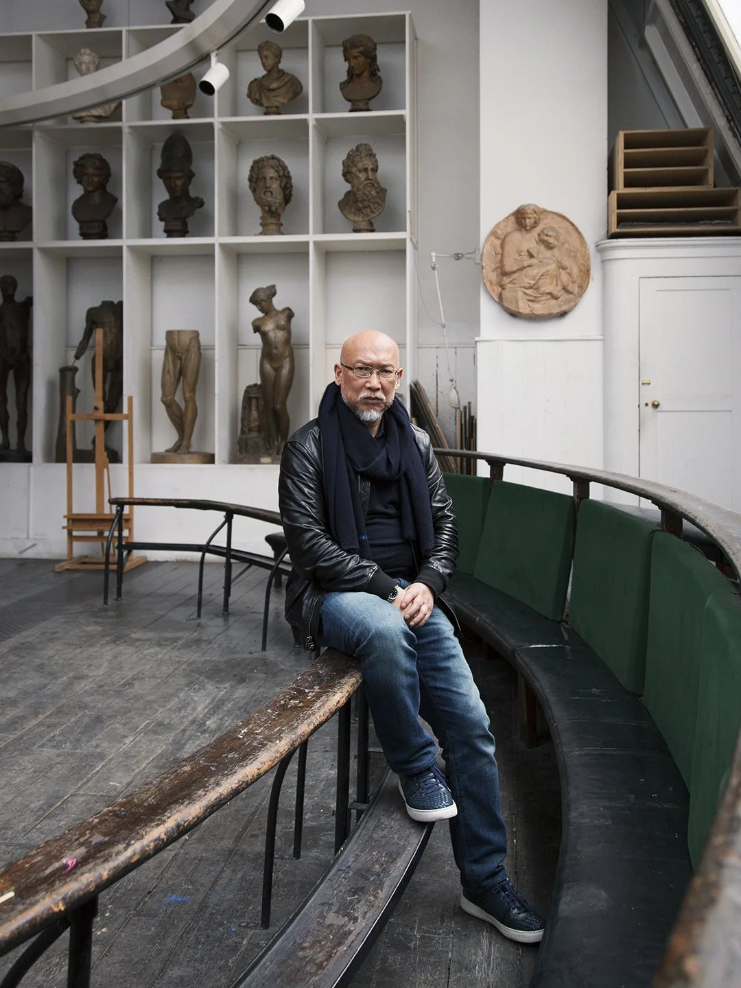 A man wearing glasses, a black leather jacket, and blue jeans sits on a wooden bench inside an art studio or museum, with bust sculptures and classical art pieces displayed on shelves and walls behind him.