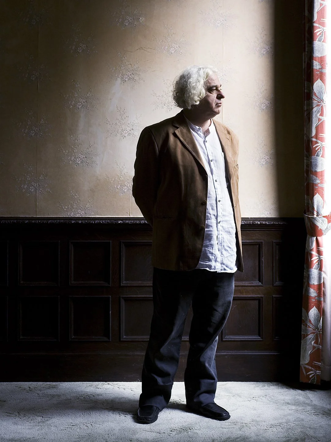 A man with white curly hair stands indoors, facing sideways near a window with light shining on him, against a decorated beige wall with dark wood paneling and floral curtains.