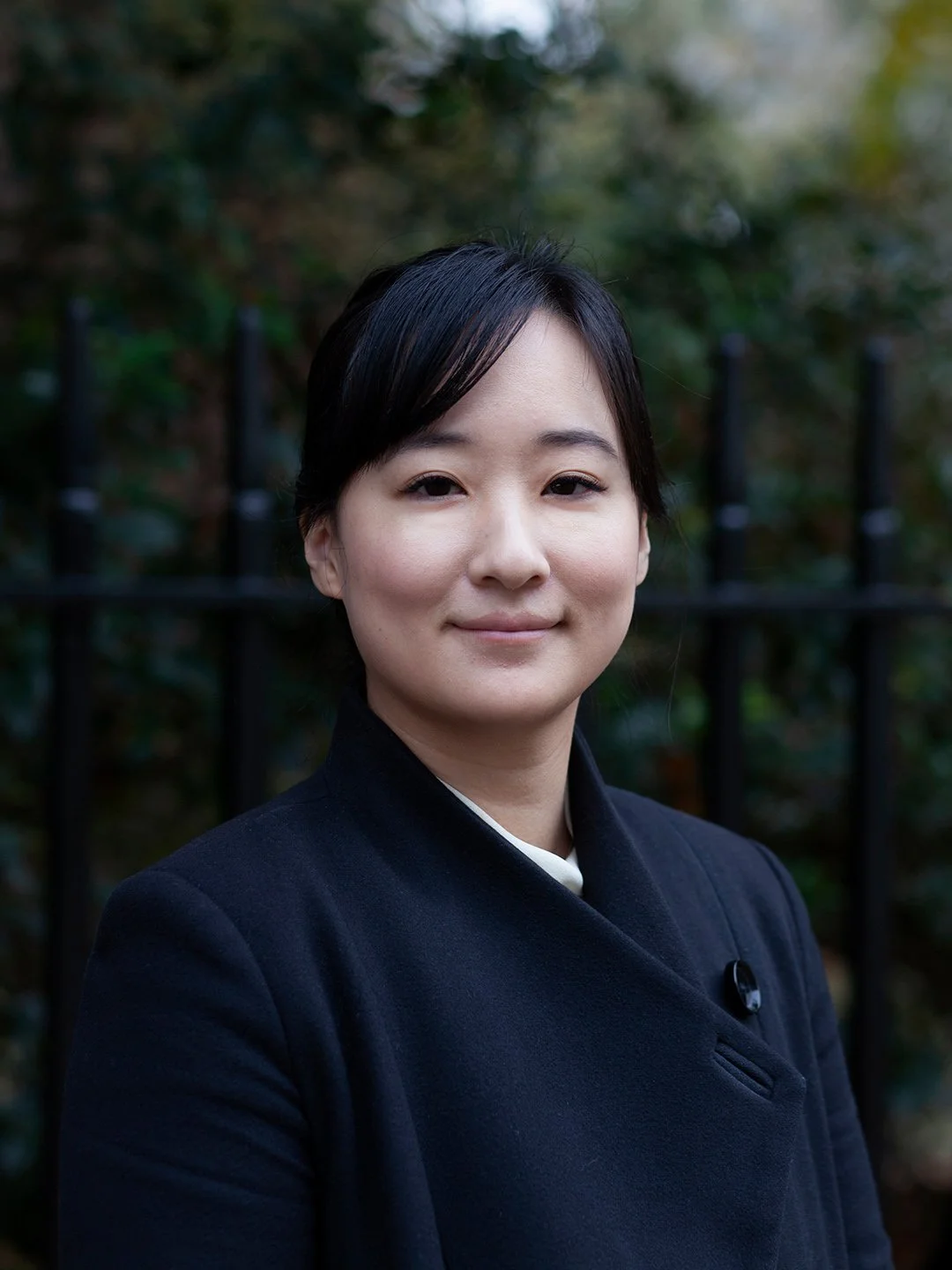 A young woman with short black hair, wearing a dark blazer, standing outdoors in front of a black fence with blurred trees and foliage in the background.