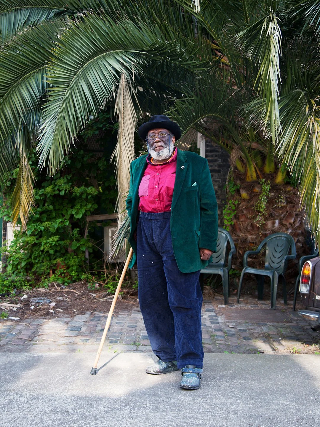 An elderly man with a beard and glasses standing outdoors in front of palm trees, wearing a black hat, green blazer, red shirt, blue pants, and holding a walking cane.